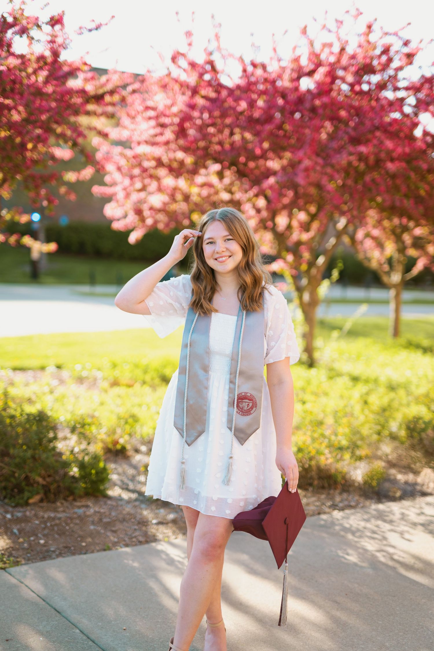 A graduate in white dress poses under blooming cherry blossom trees with graduation stole.
