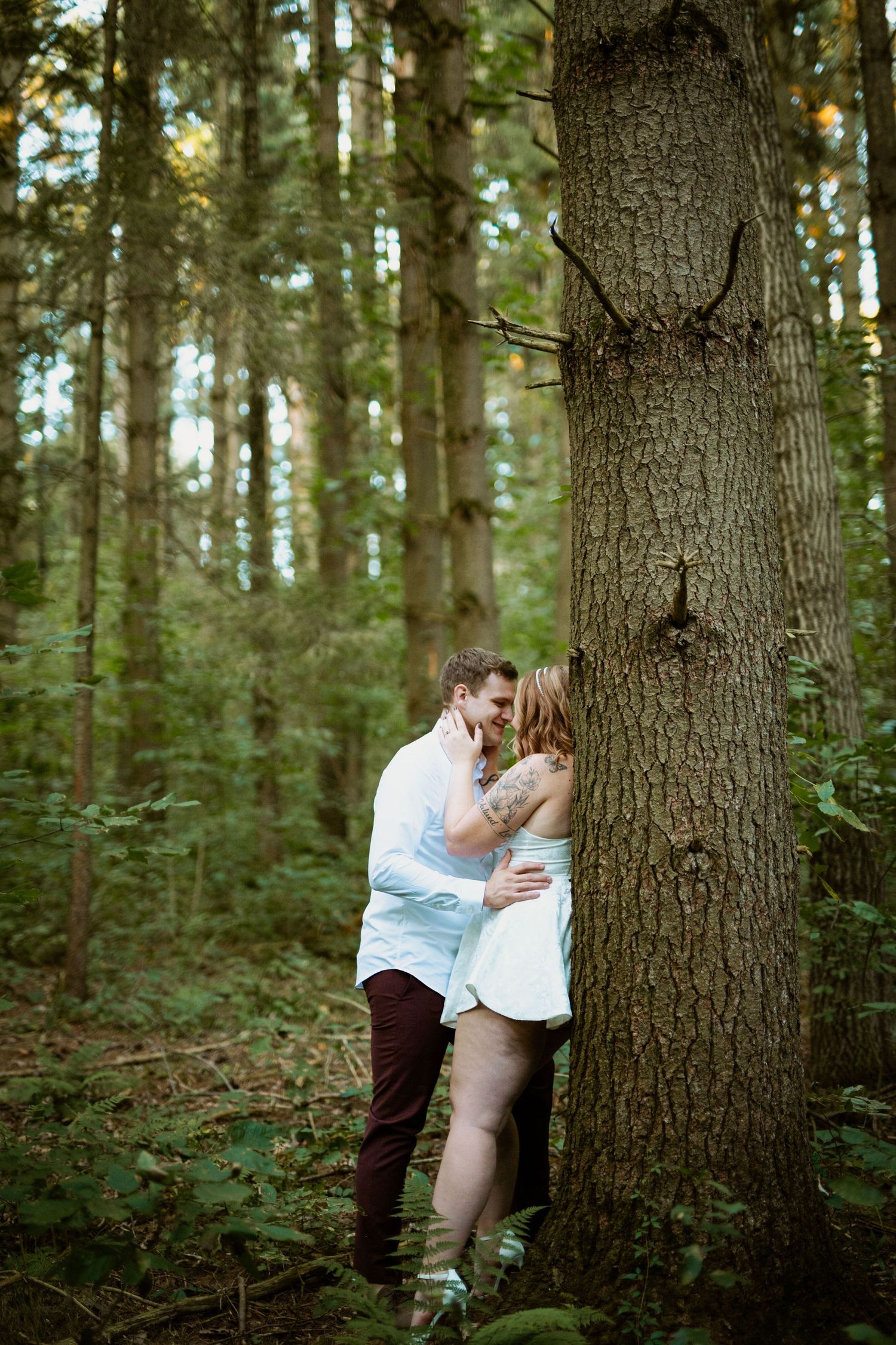 A romantic moment captured between a couple by a large tree trunk in a forest setting.