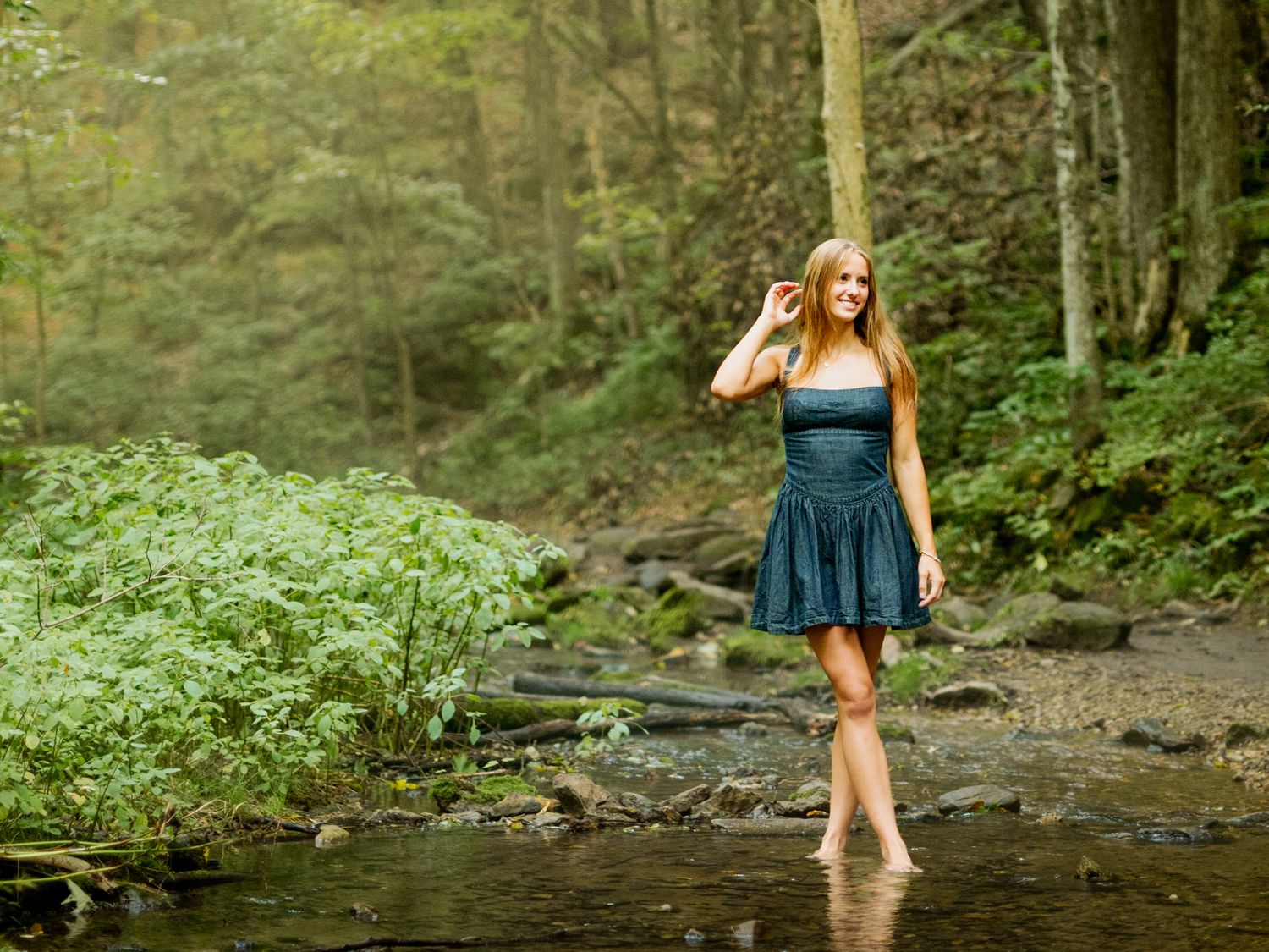 A person in a navy blue dress poses near a forest stream during a summer photoshoot.