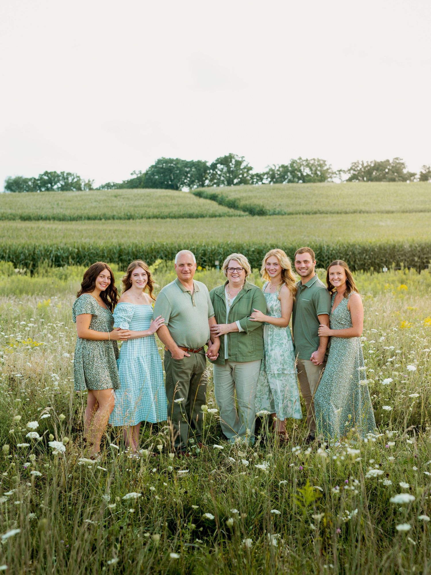 A group in matching sage green and mint dresses poses together in a wildflower field during golden hour.