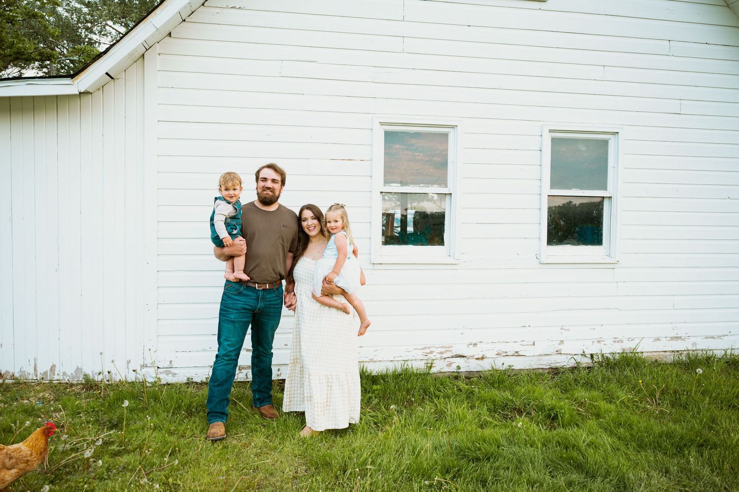 A group of people stand together on grass in front of a white building with two windows.