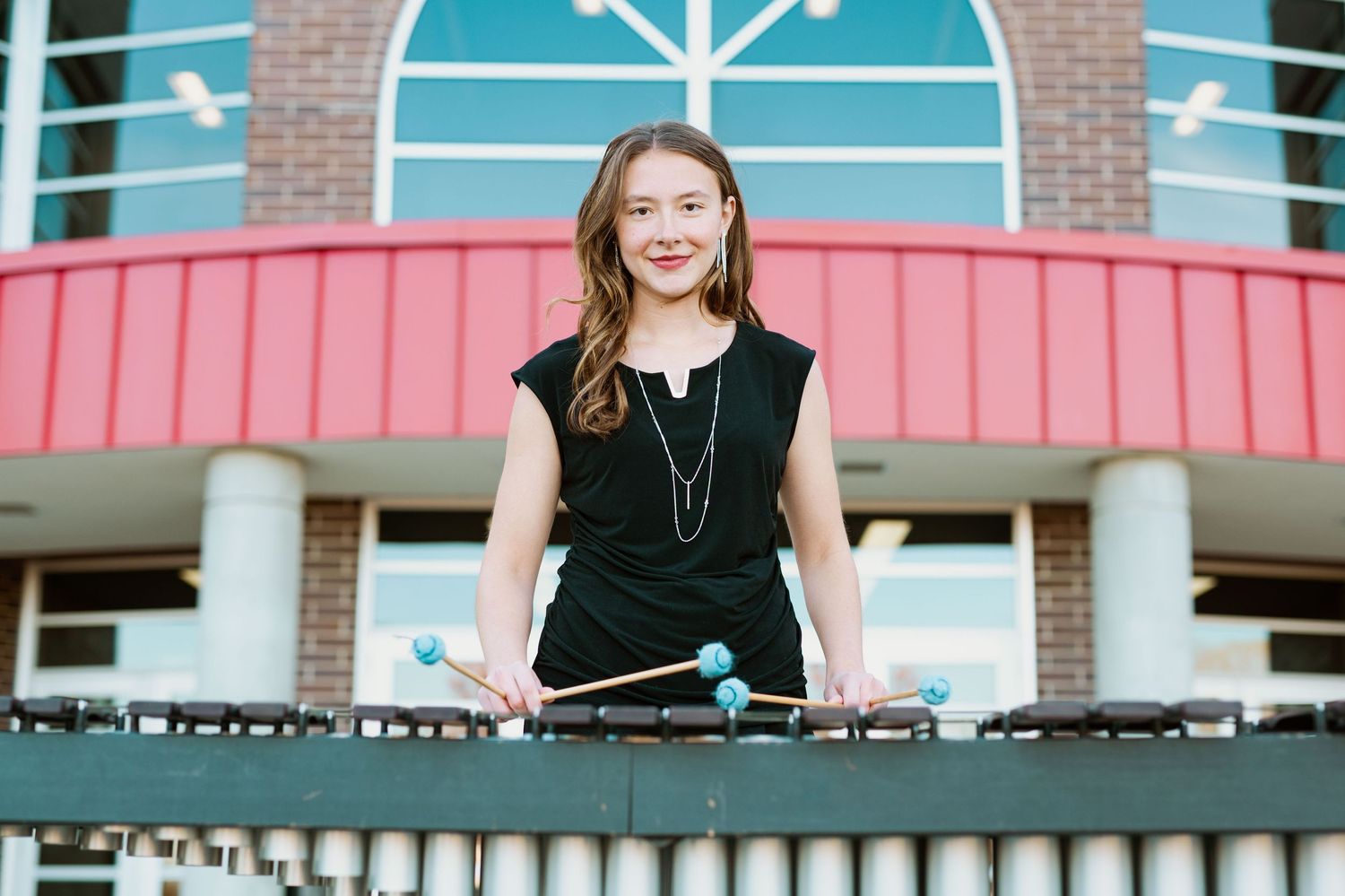 Band member poses with mallets at vibraphone in front of brick building entrance.
