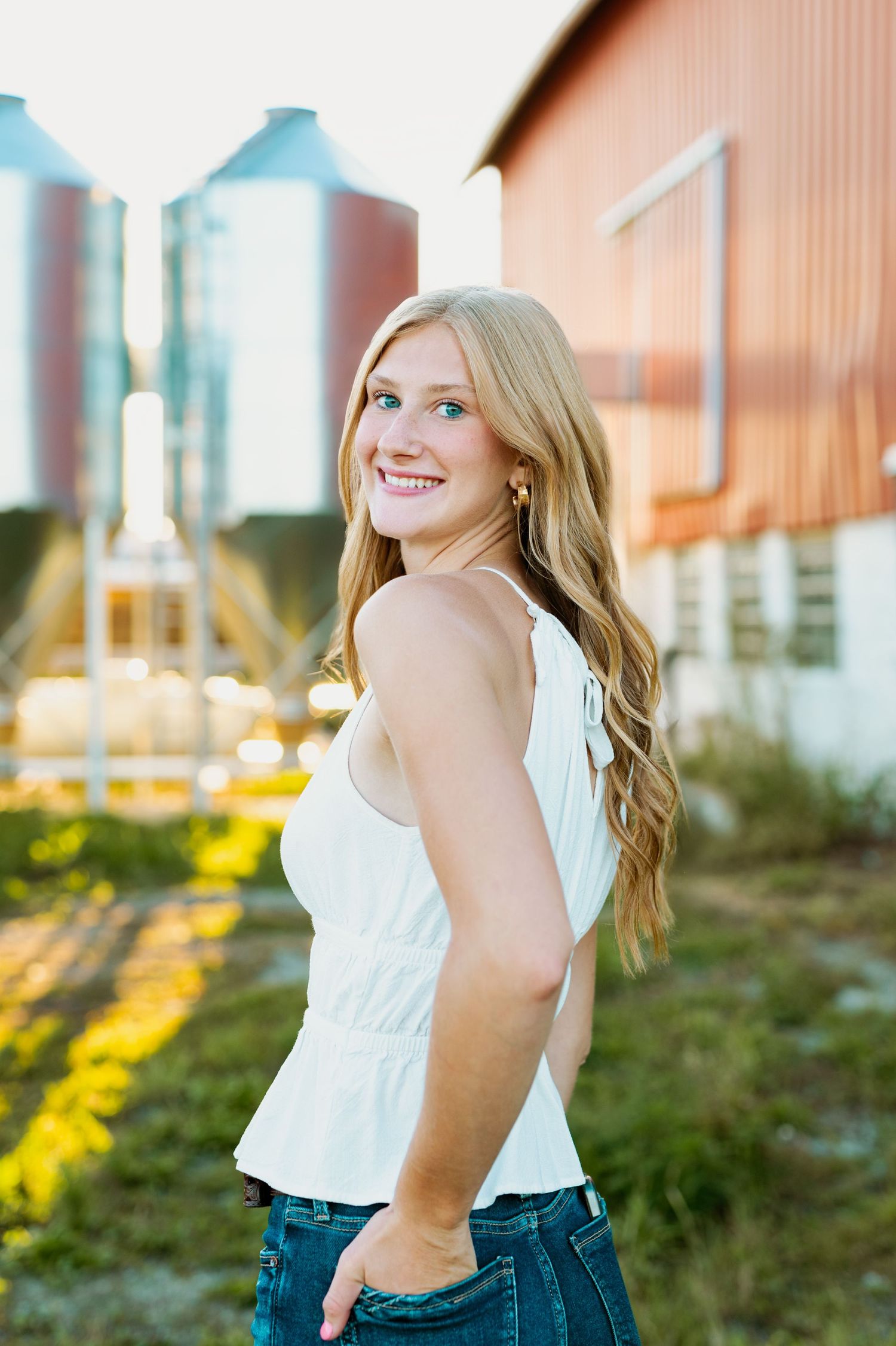 Portrait of a person in a white sleeveless top and jeans posing outdoors near farm silos at golden hour.
