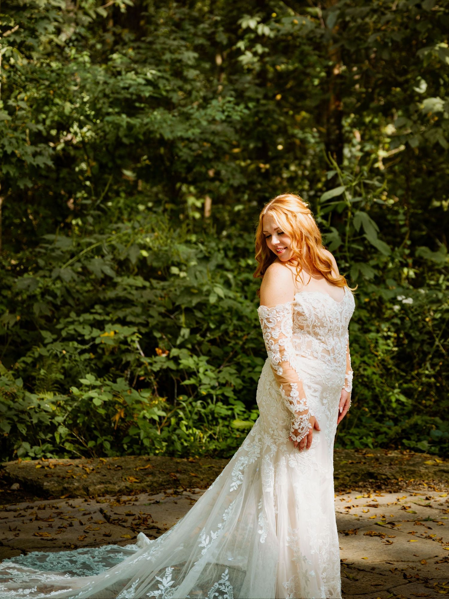 A couple in wedding attire embraces on a forest path with trailing lace dress details.