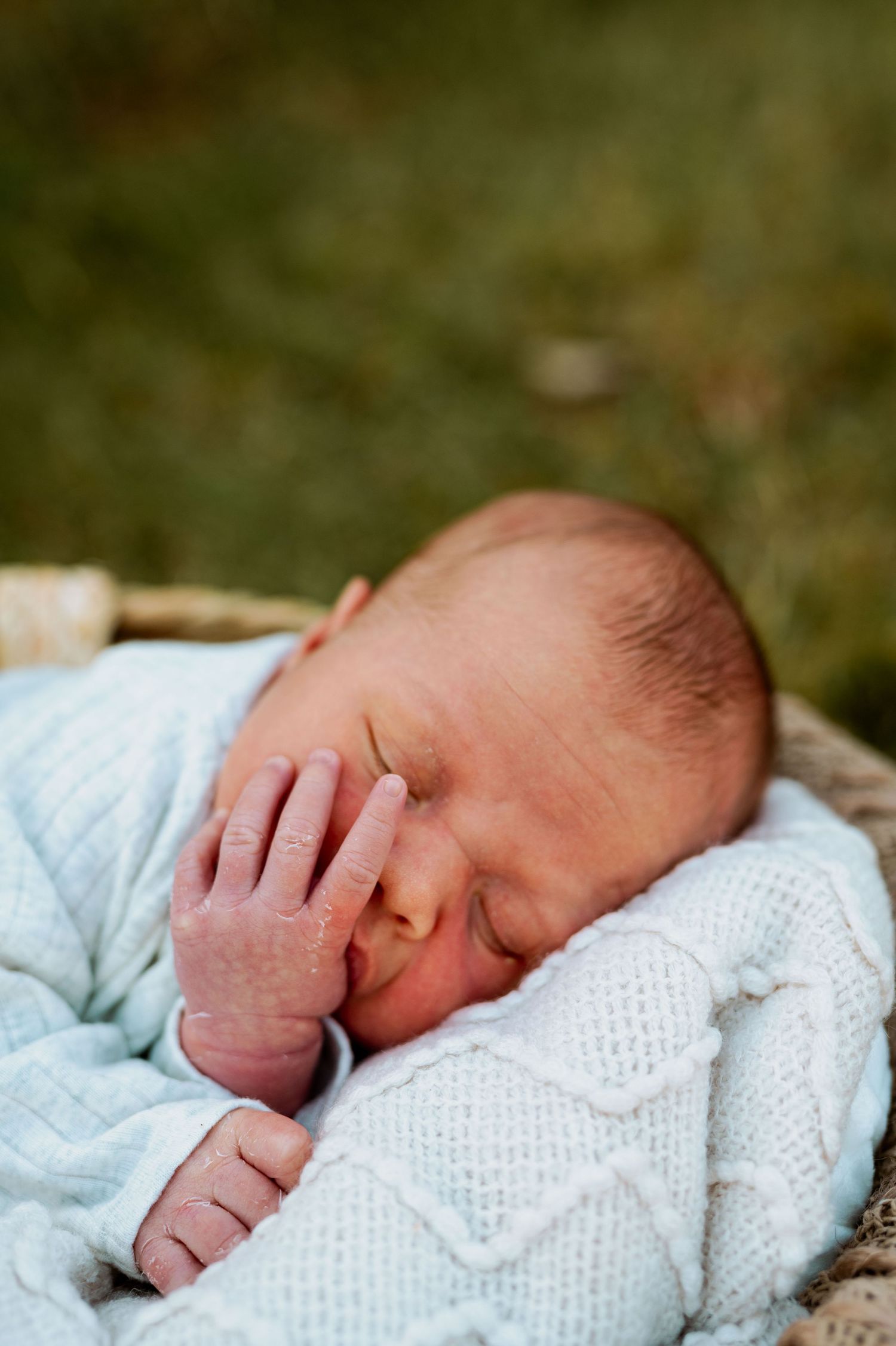 A sleeping newborn baby wrapped in a soft white knitted blanket rests peacefully in a natural outdoor setting.