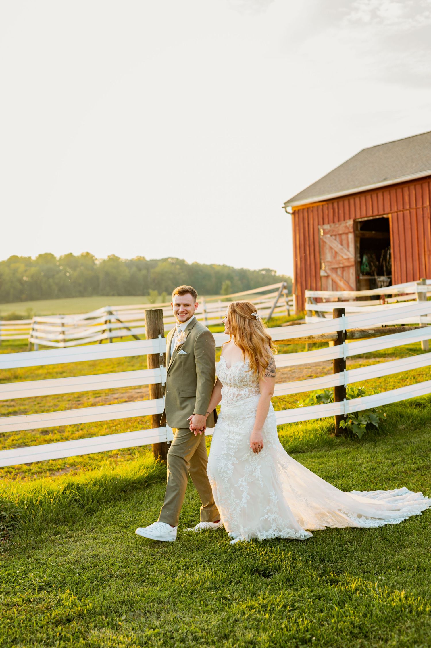 Newlyweds walking near a rustic red barn and white fence during golden hour.