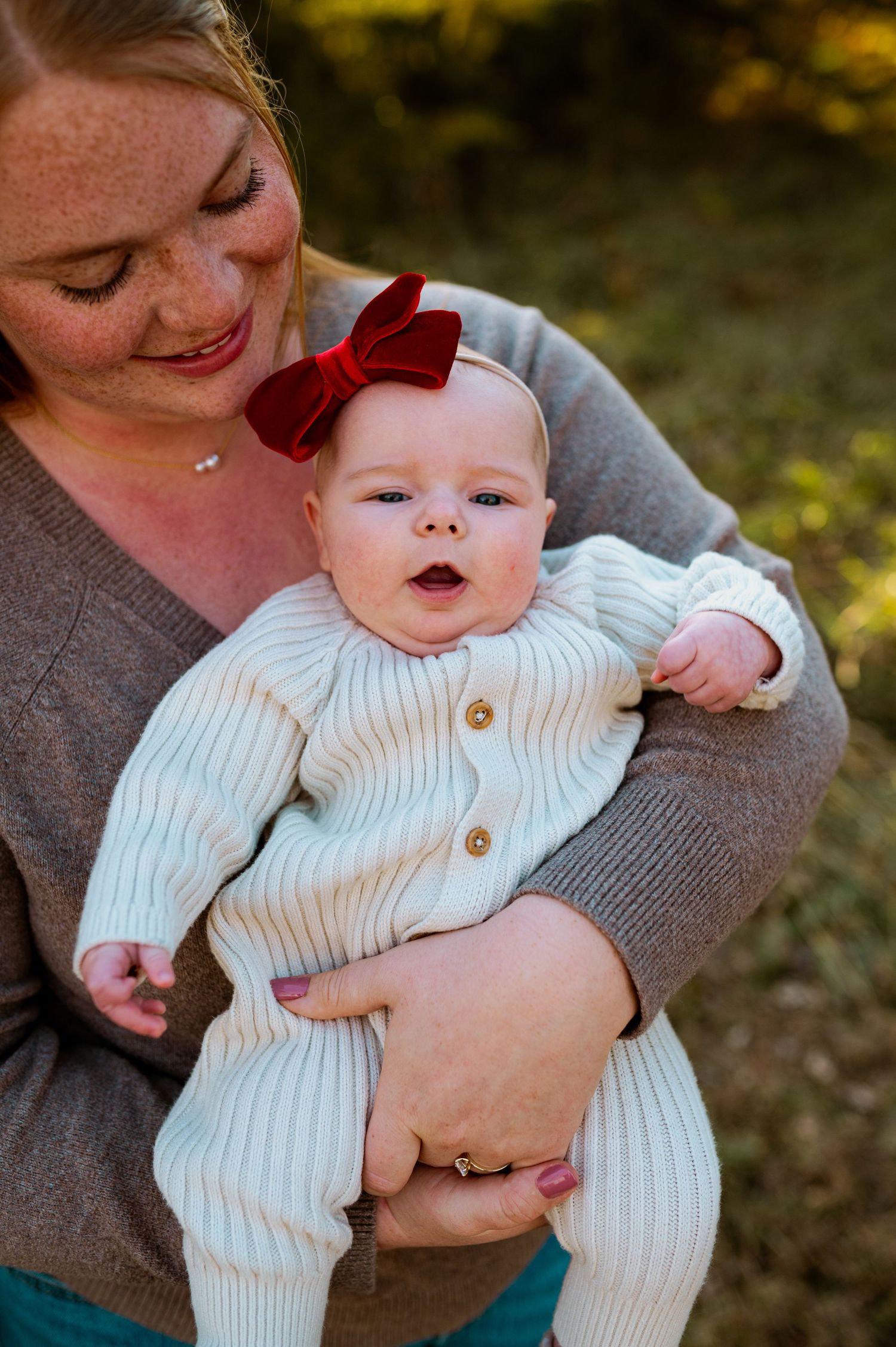 A parent in a gray sweater holds and snuggles with a baby wearing a white knit sweater outdoors during autumn.