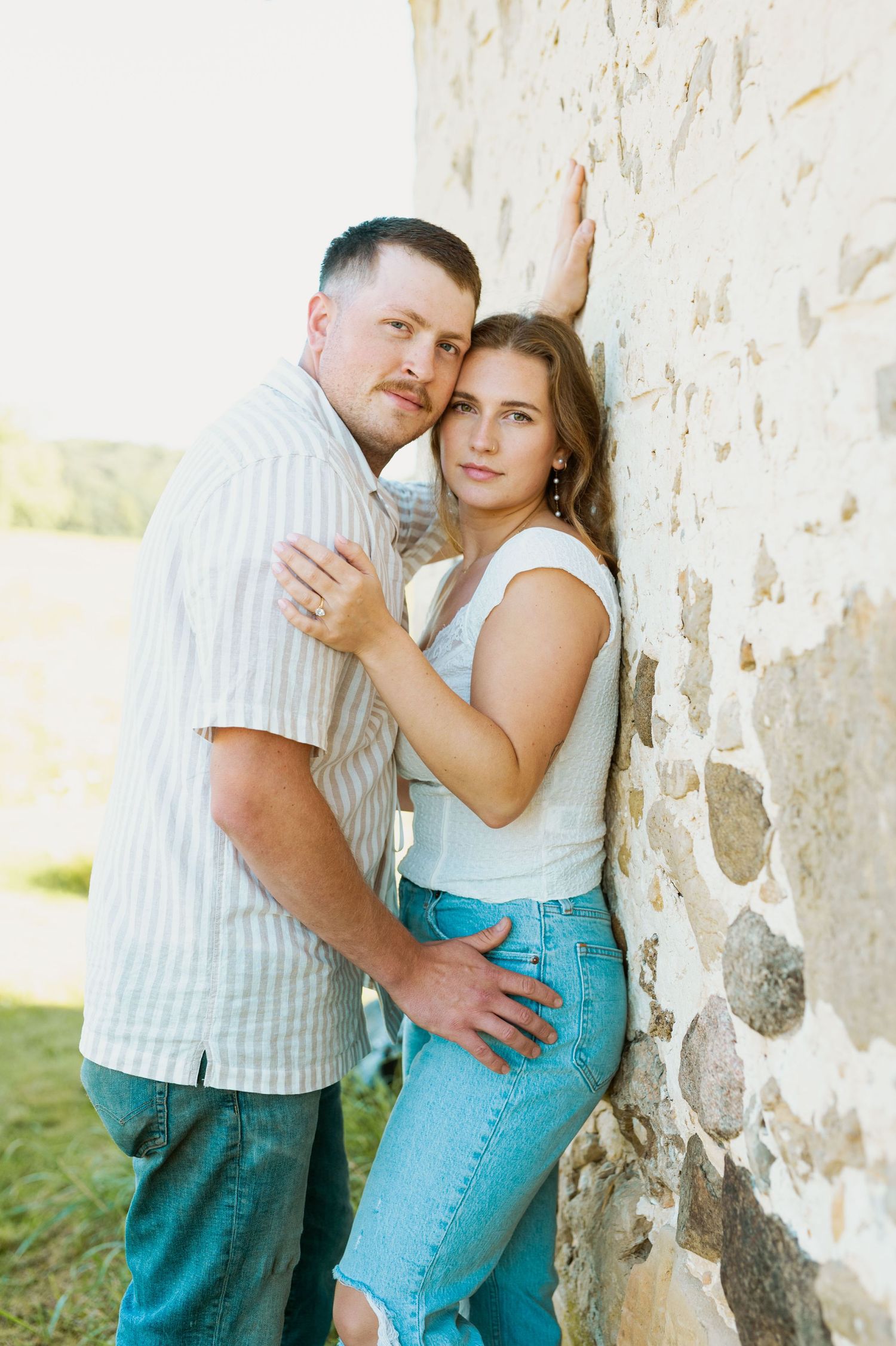 A romantic couple pose against a weathered stone wall outdoors.