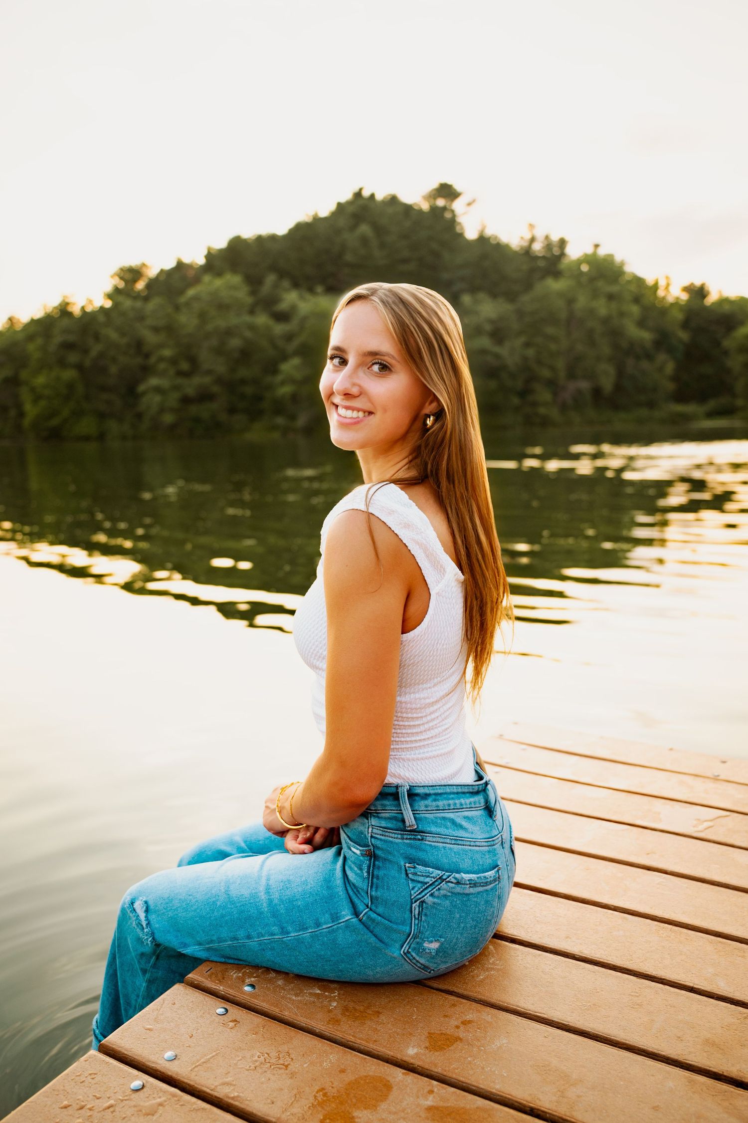 Person in blue jeans and white top sits on wooden dock by lake at sunset.