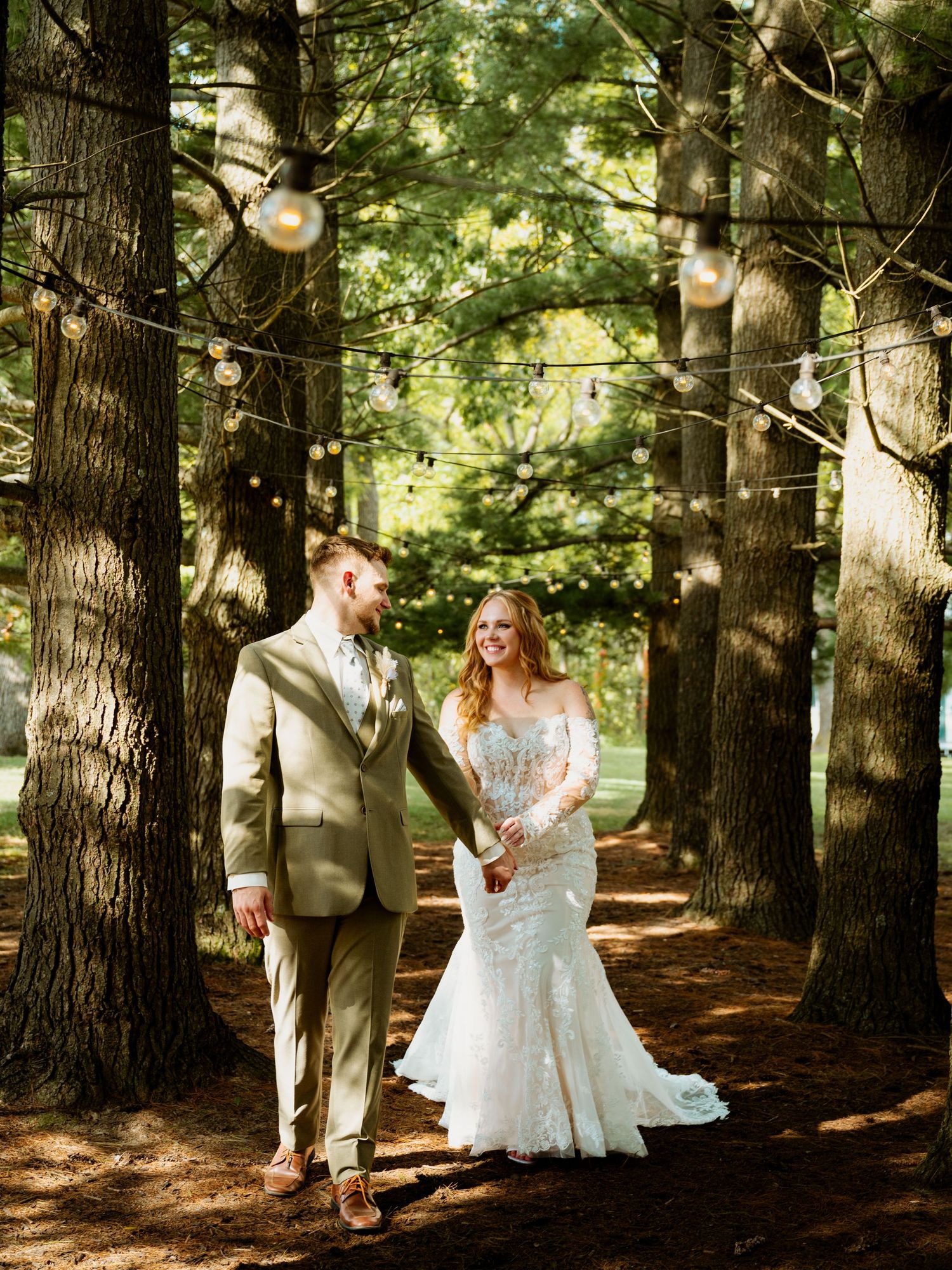 A couple walks hand in hand through a dreamy forest path with string lights hanging between tall pine trees.