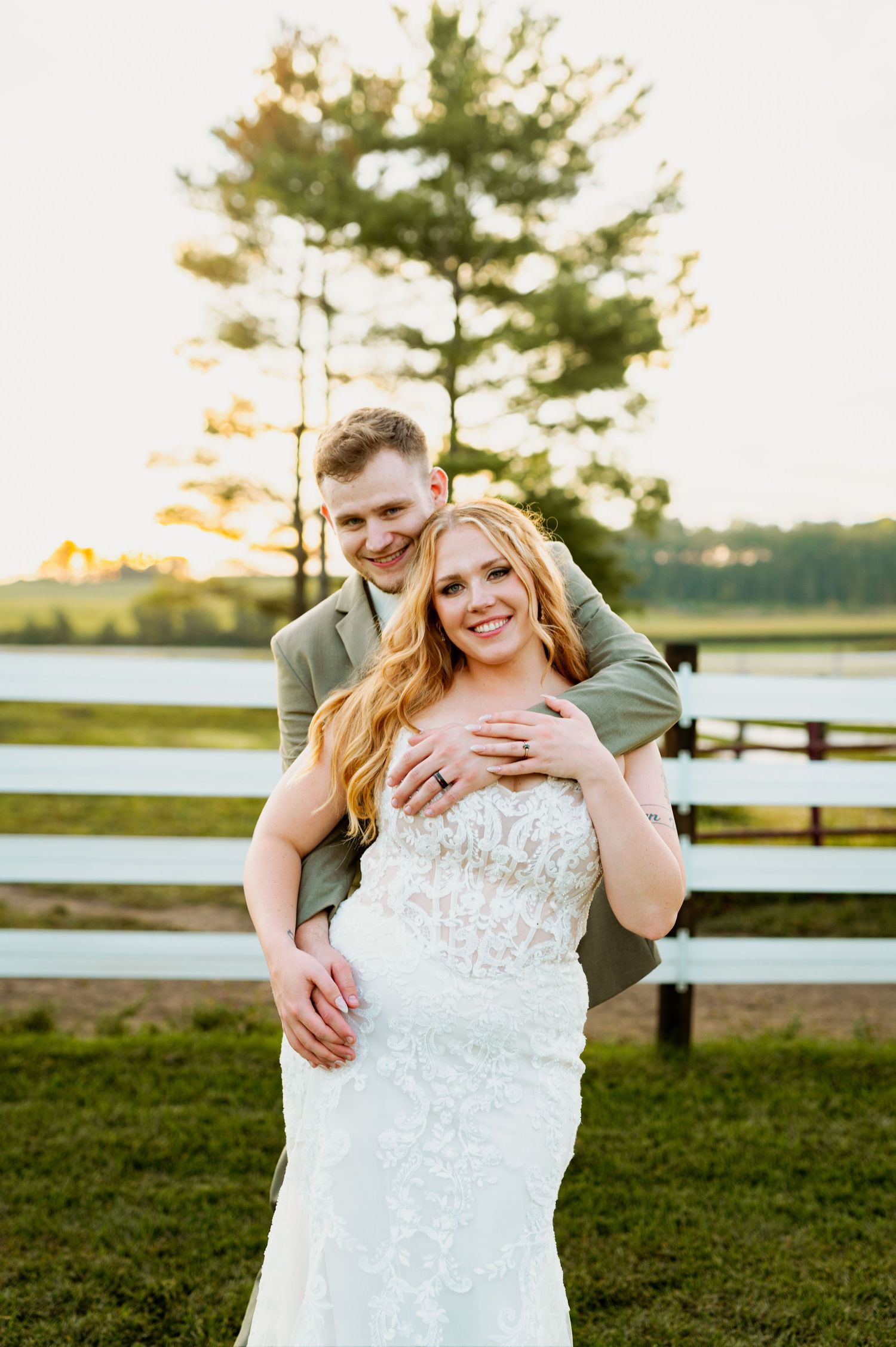 A couple shares a romantic sunset moment outdoors with a pine tree and wooden fence in the background.