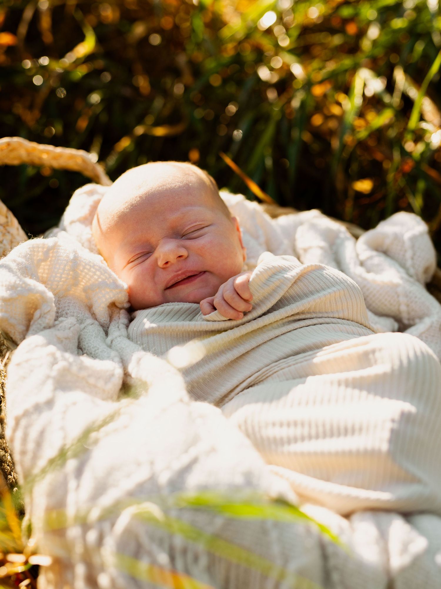 A sleeping baby wrapped in a cream-colored knit blanket rests peacefully in natural outdoor lighting.