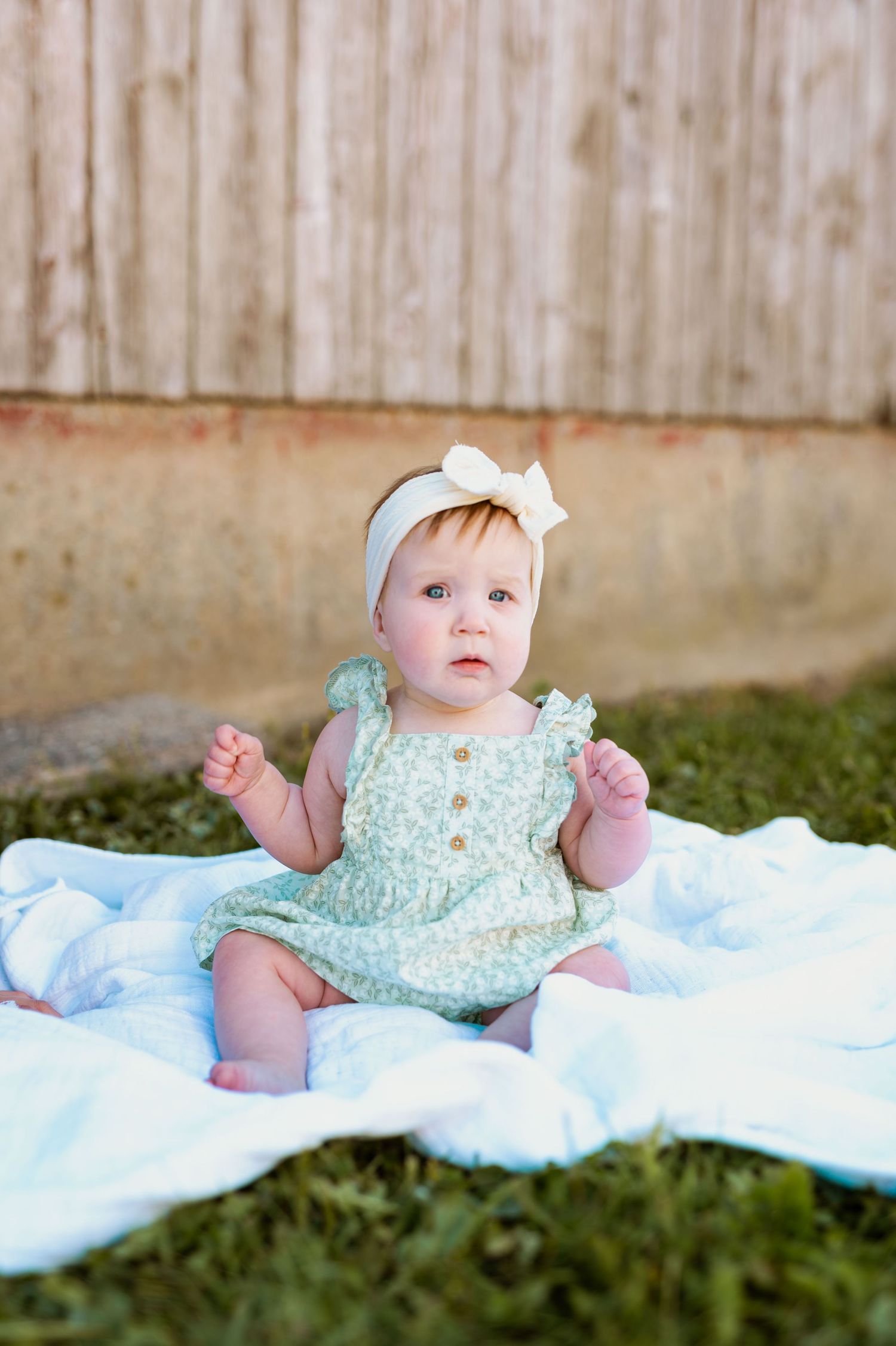 Baby wearing mint green dress and white headband sits on blanket against wooden fence.