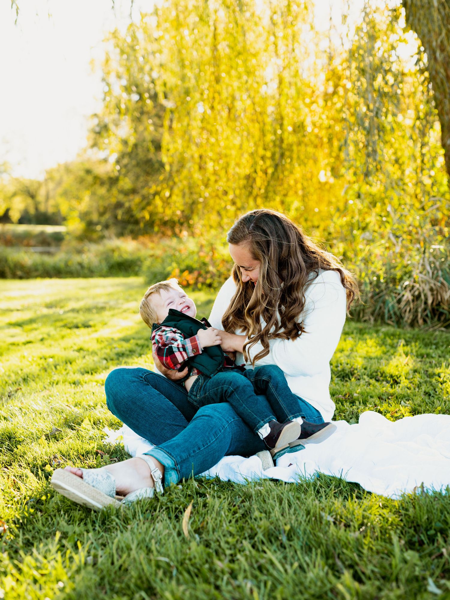 Person in teal pants relaxing on white blanket with camera in sunny autumn field with golden foliage.