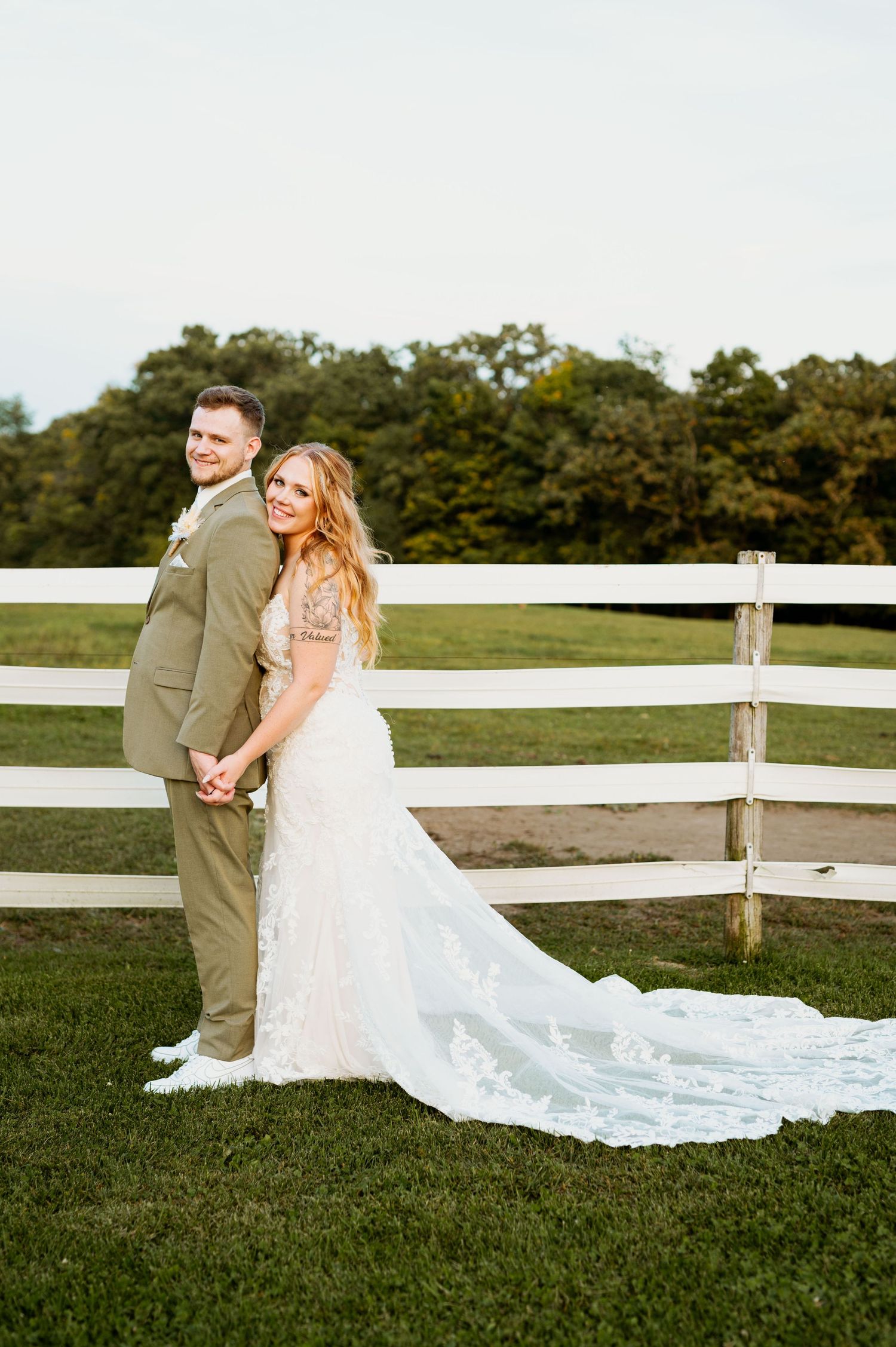 Elegant wedding couple poses by white fence with flowing lace train spread across green grass.