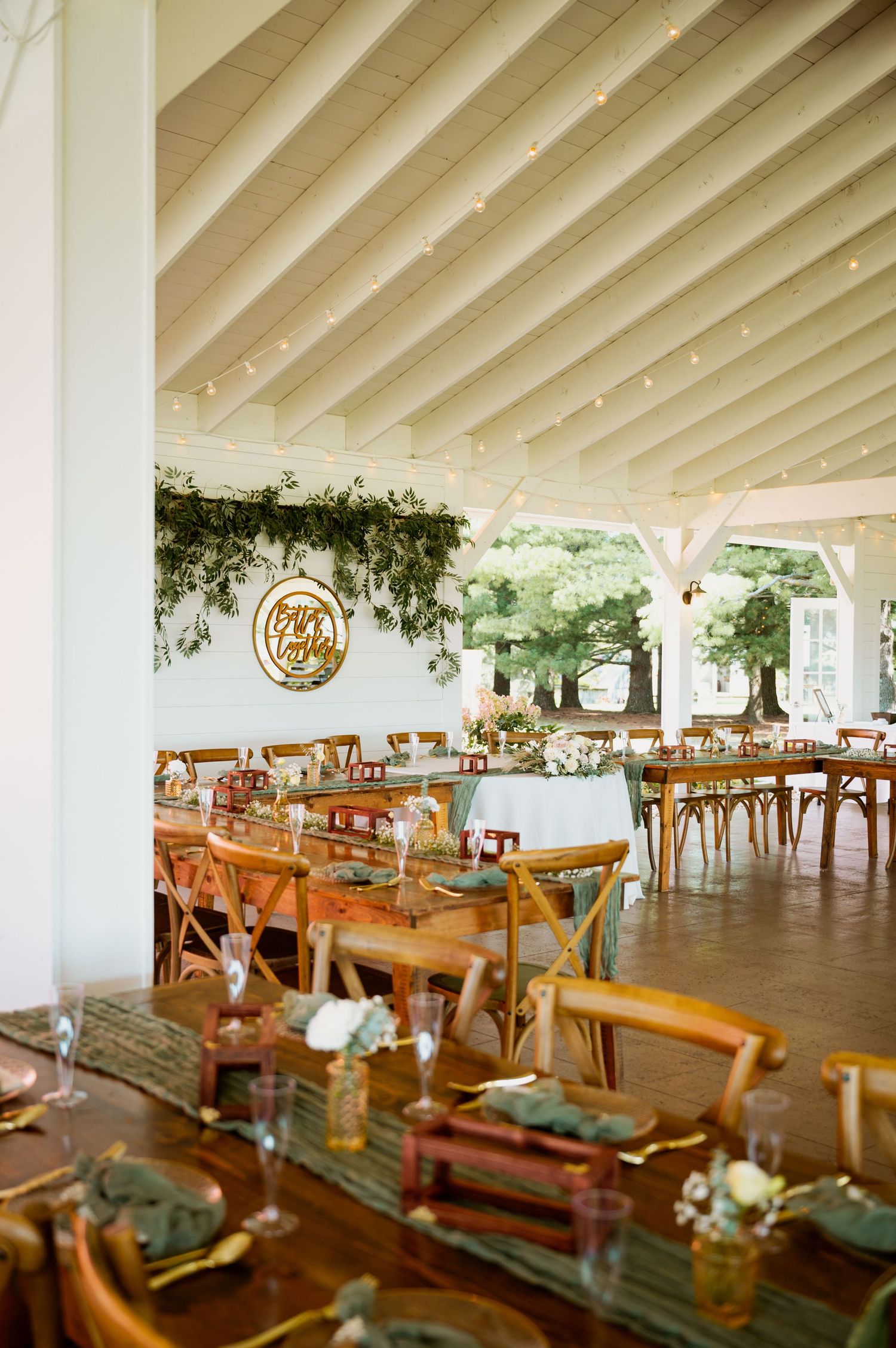 Rustic wedding reception venue with wooden tables and chairs arranged under a white beamed ceiling with hanging decorations.