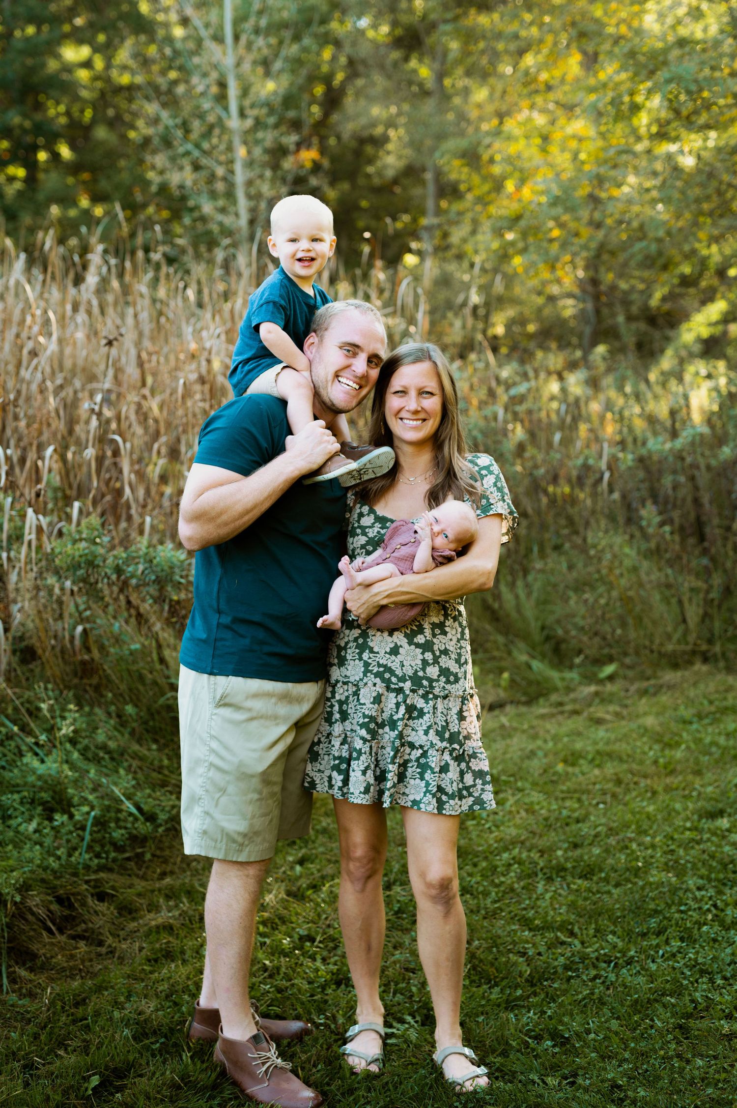 A family poses together outdoors in a natural setting with tall grass and autumn foliage in the background.