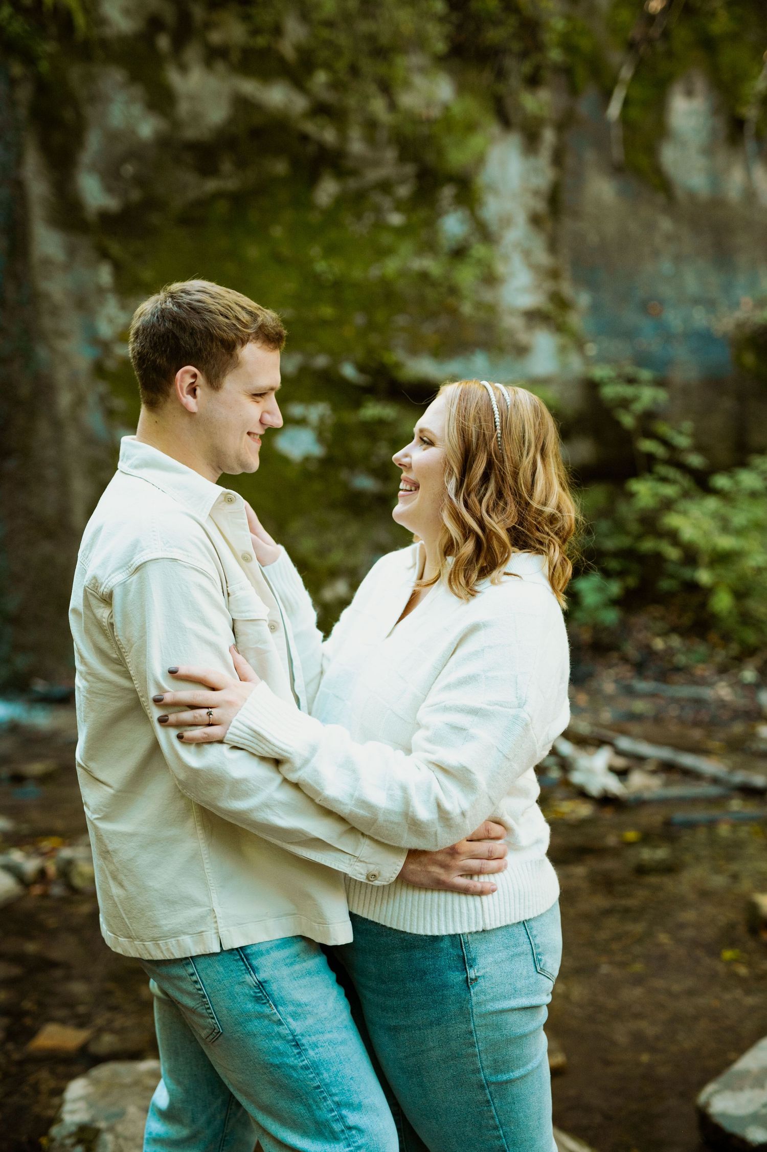 A couple wearing matching cream sweaters embrace during an outdoor photo shoot near mossy rocks.