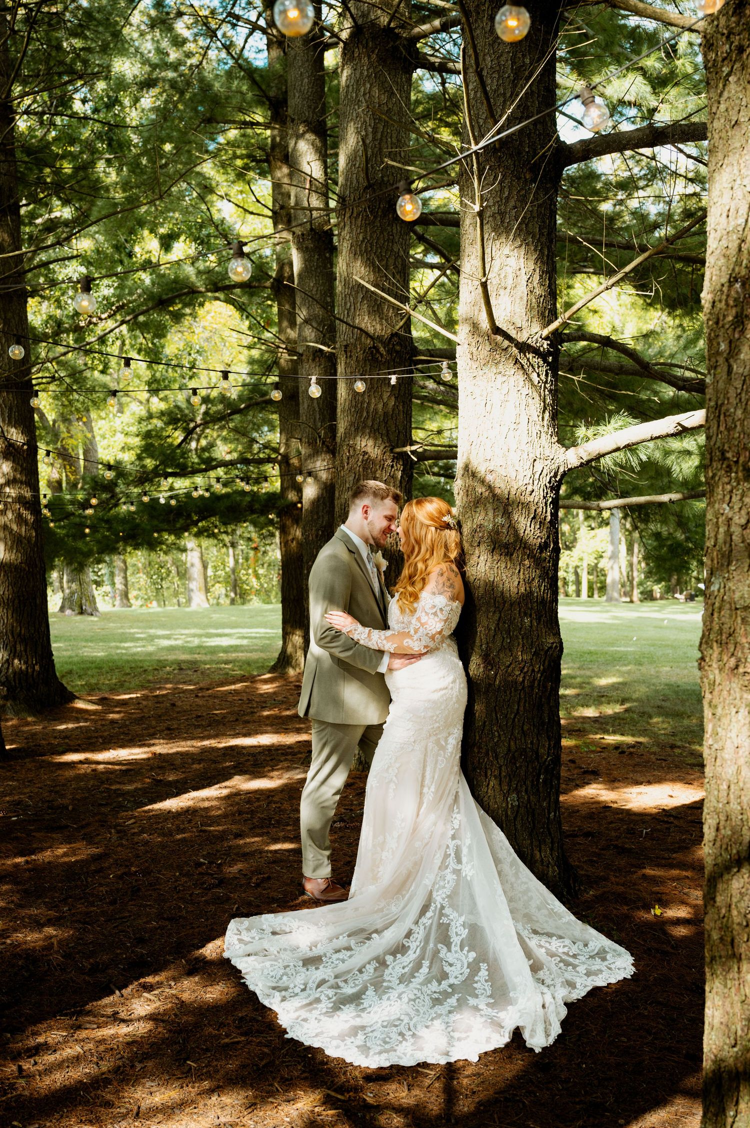 A romantic wedding photo in a forest setting with string lights overhead and a stunning lace train dress.