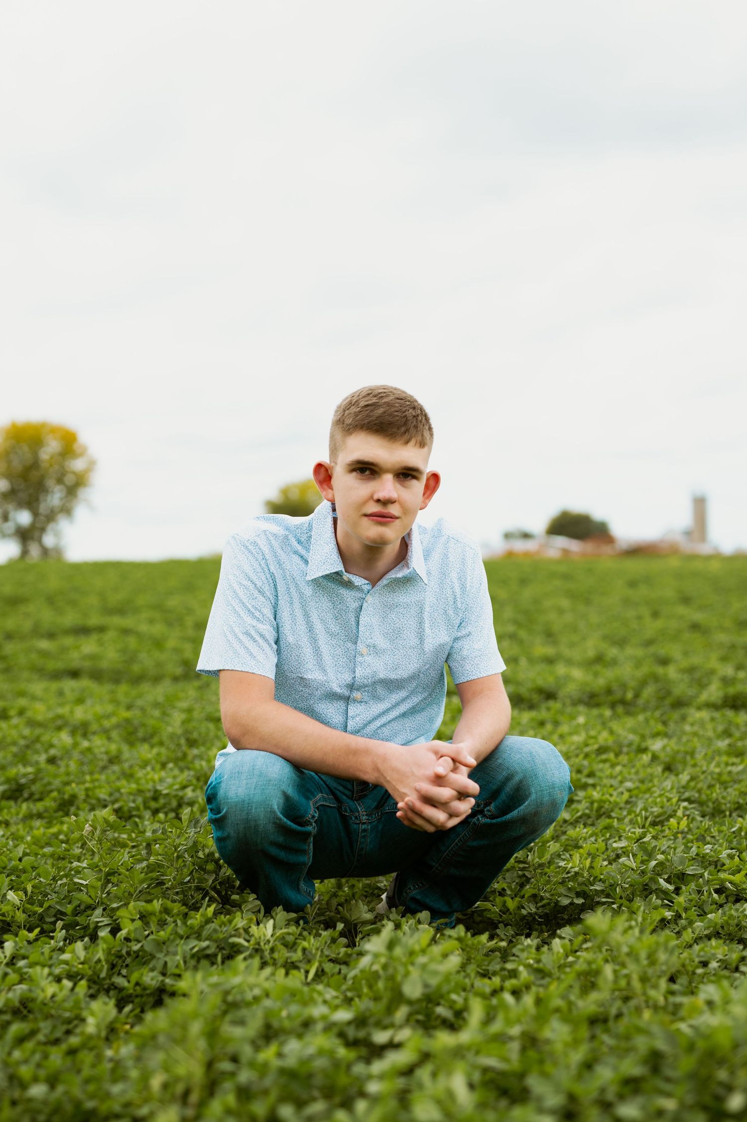 Person crouching in green field wearing light blue shirt and jeans under cloudy sky.
