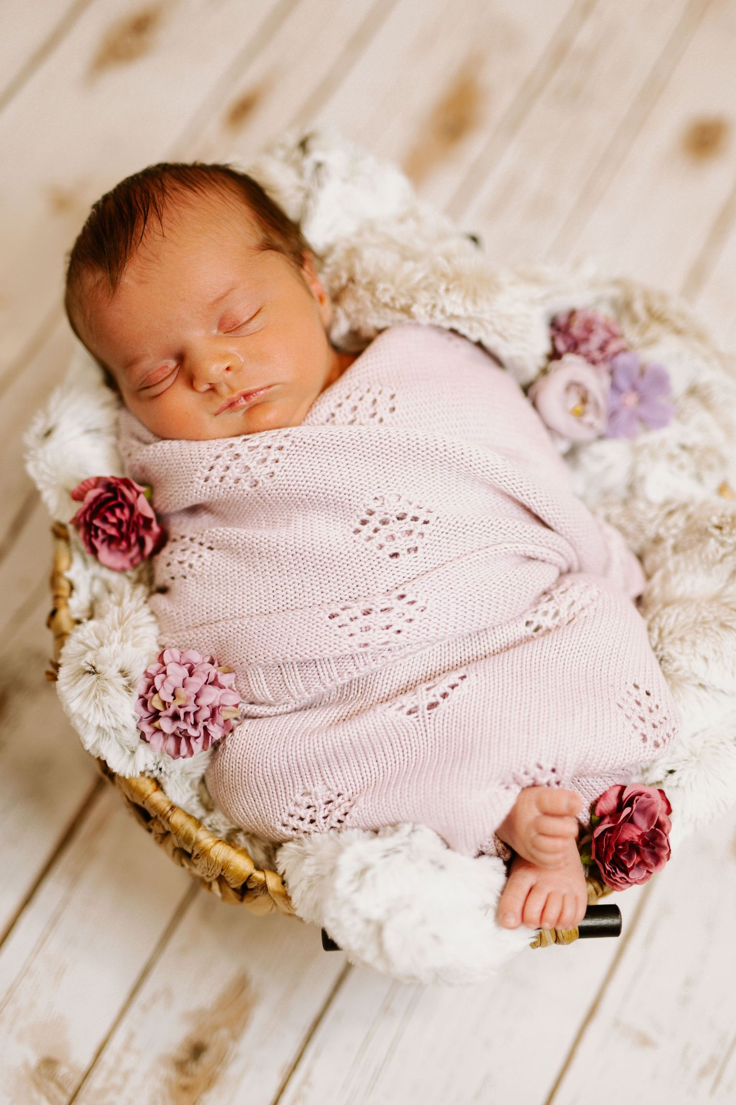 Sleeping baby in pink knit outfit with floral decorations in rustic basket.