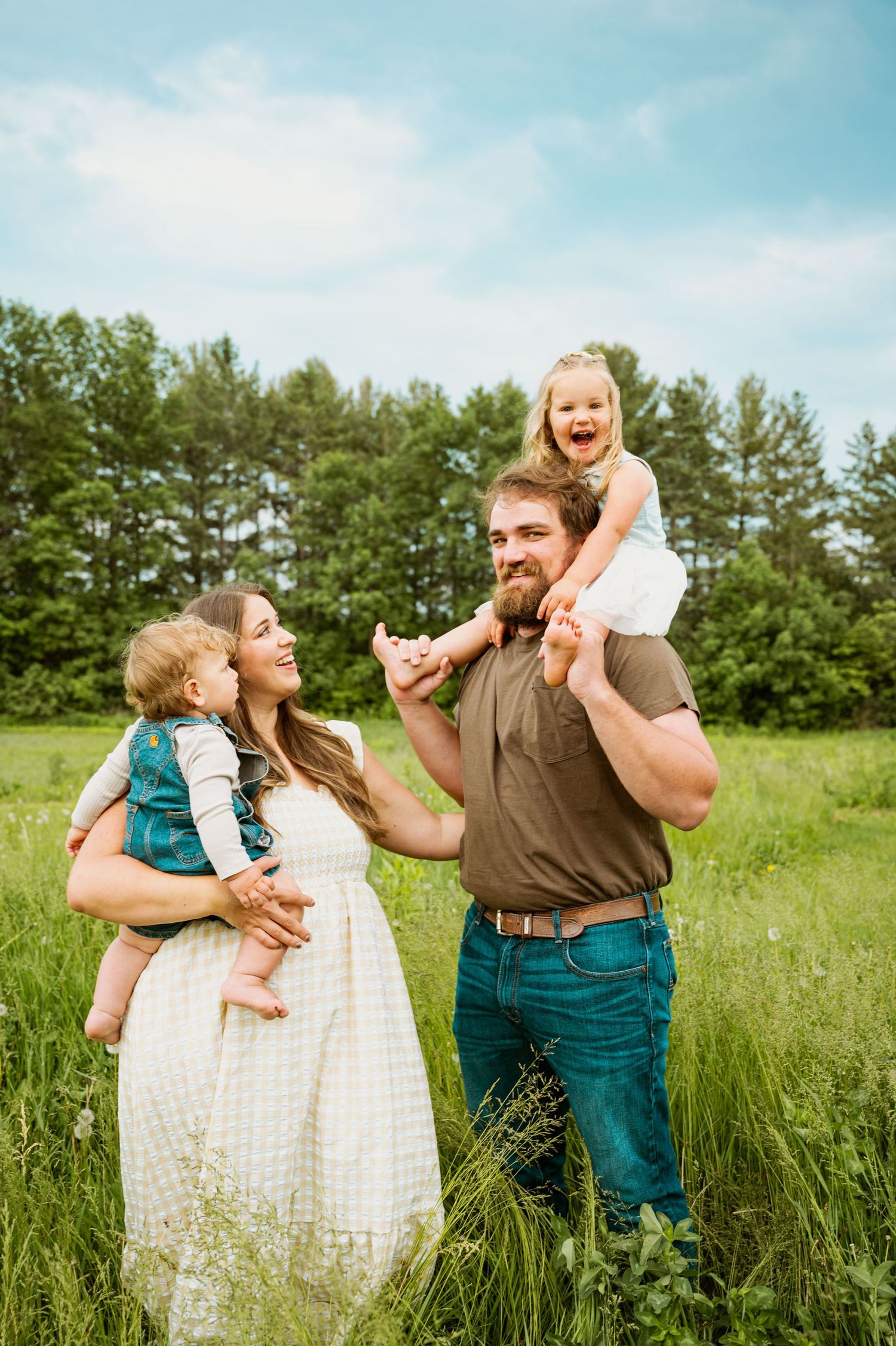 A joyful family shares laughter together in a sunny green field with pine trees in the background.