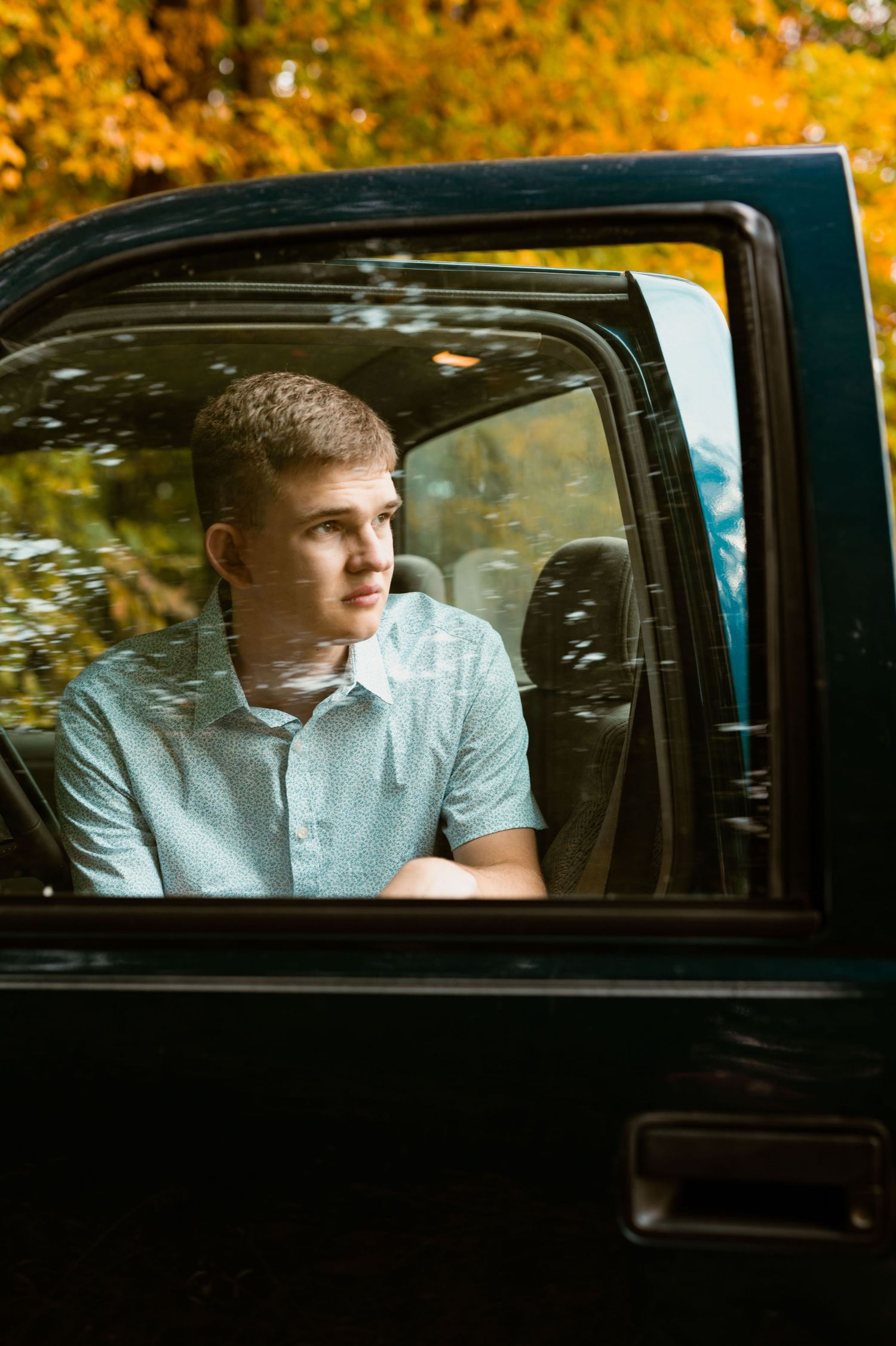 Portrait through truck window showing person in light blue shirt with autumn foliage reflection.