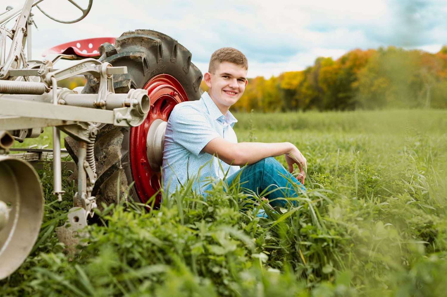 Young person in casual attire sitting in a grassy field next to vintage farm equipment during golden hour.