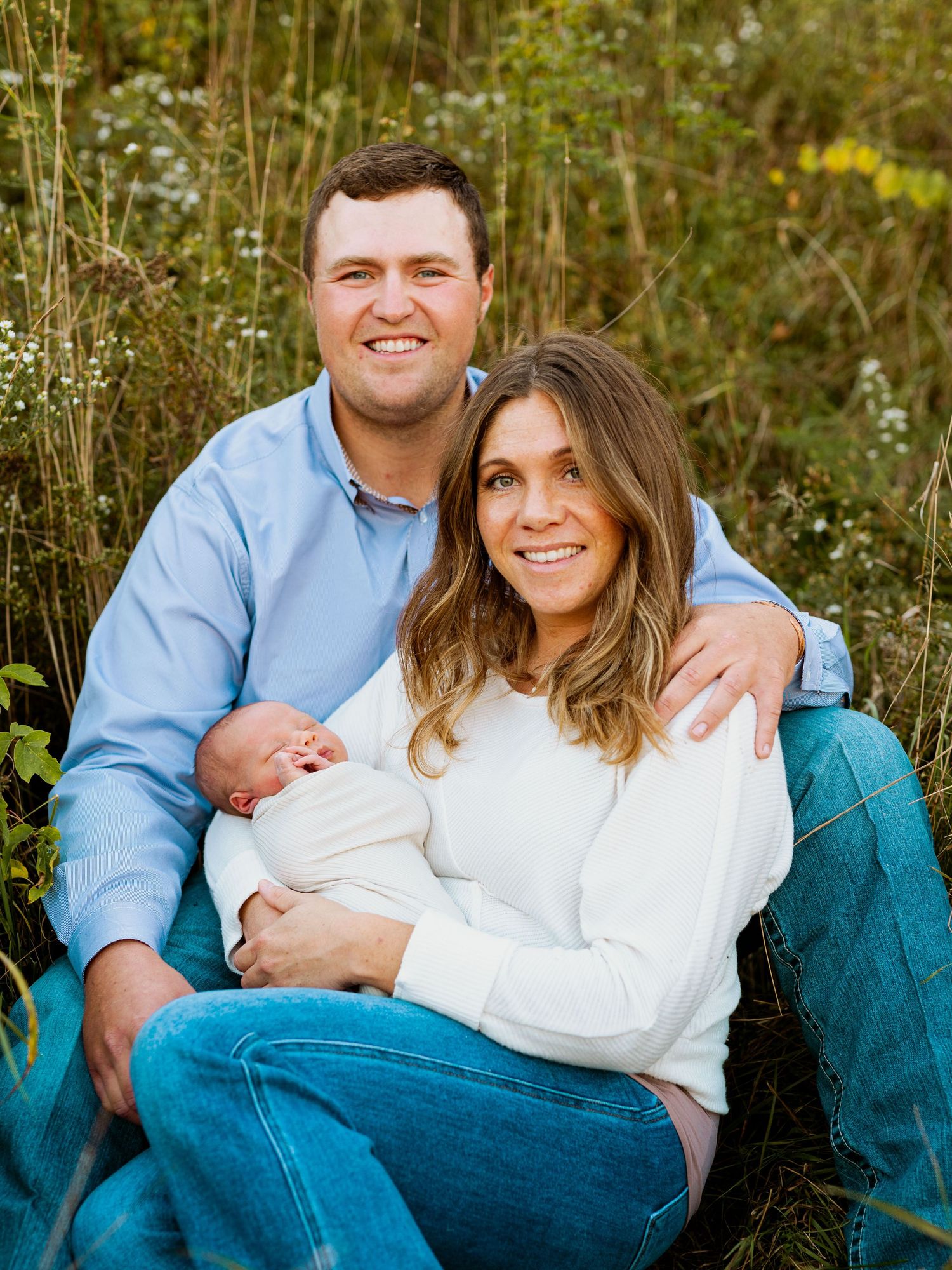A couple sits together in a teal chair outdoors surrounded by wildflowers during a family photo session.