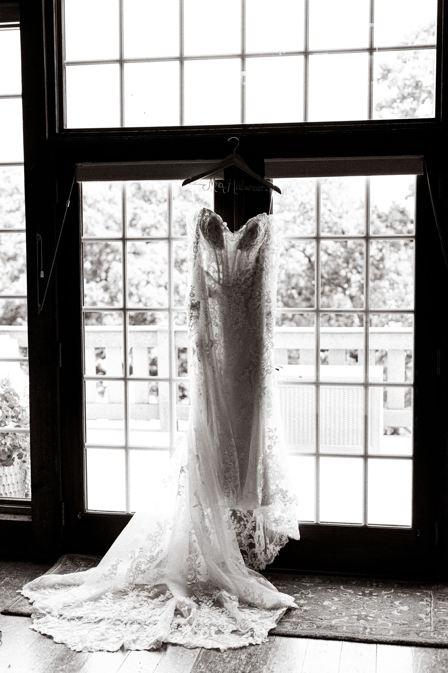 A delicate white lace wedding dress hangs elegantly in front of a bright window in black and white photography.