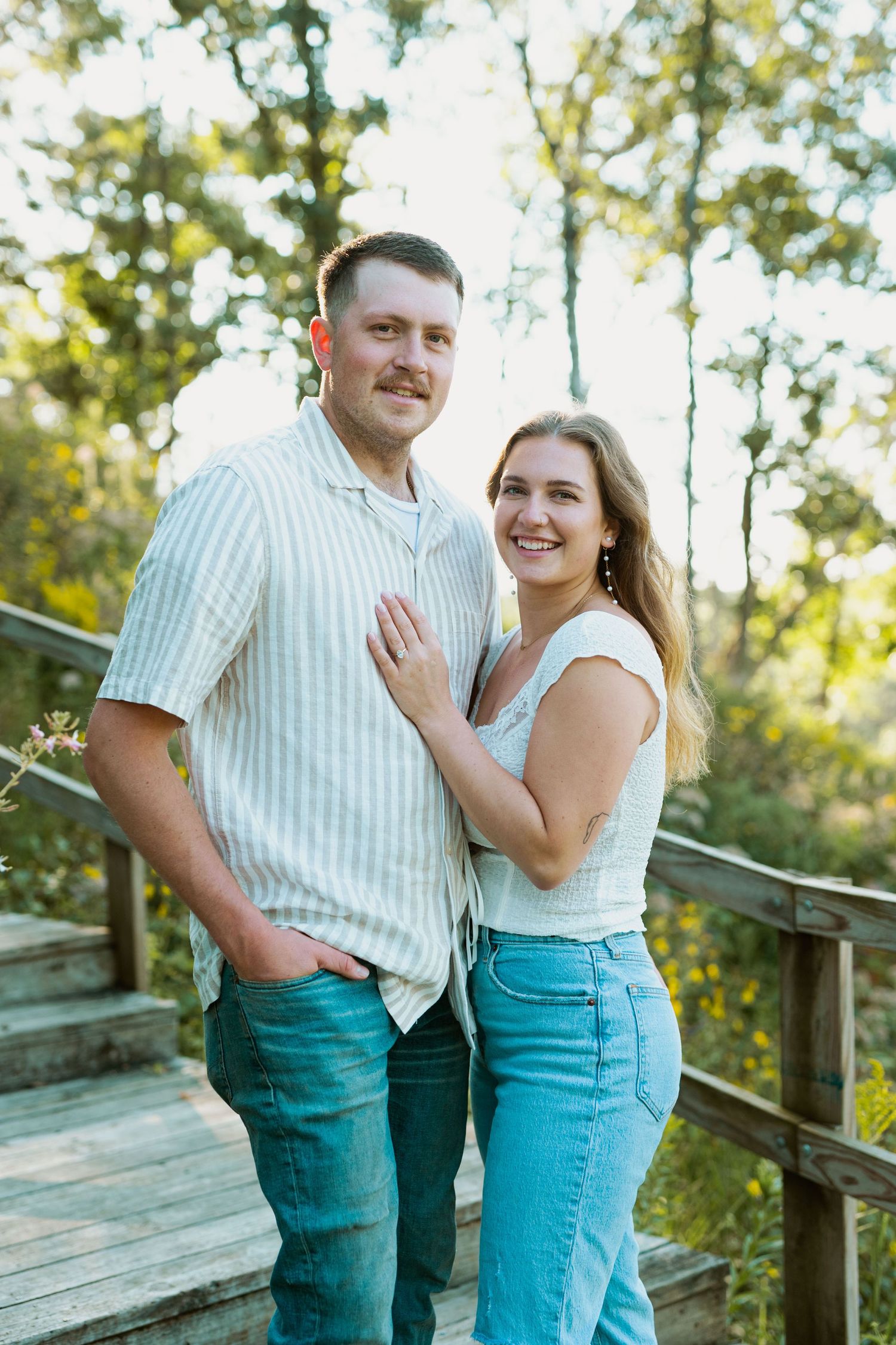 A couple embracing on a wooden deck with trees and natural lighting in the background.