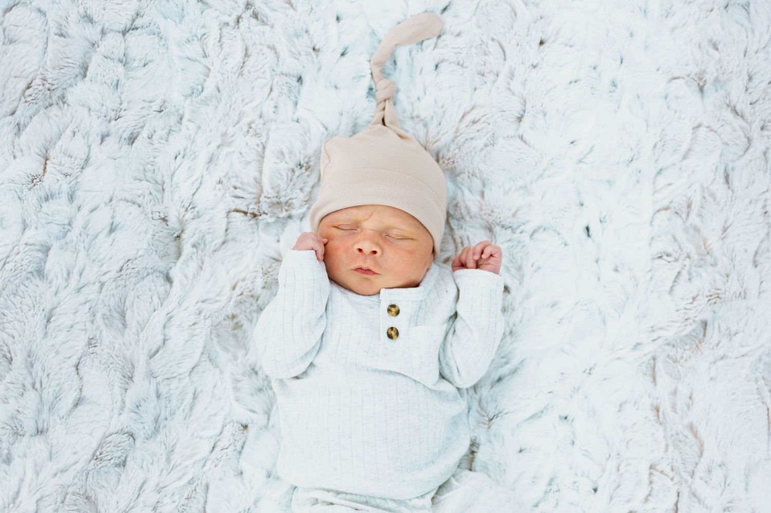 A newborn baby wearing a white knit hat lies peacefully on soft marble-patterned bedding.