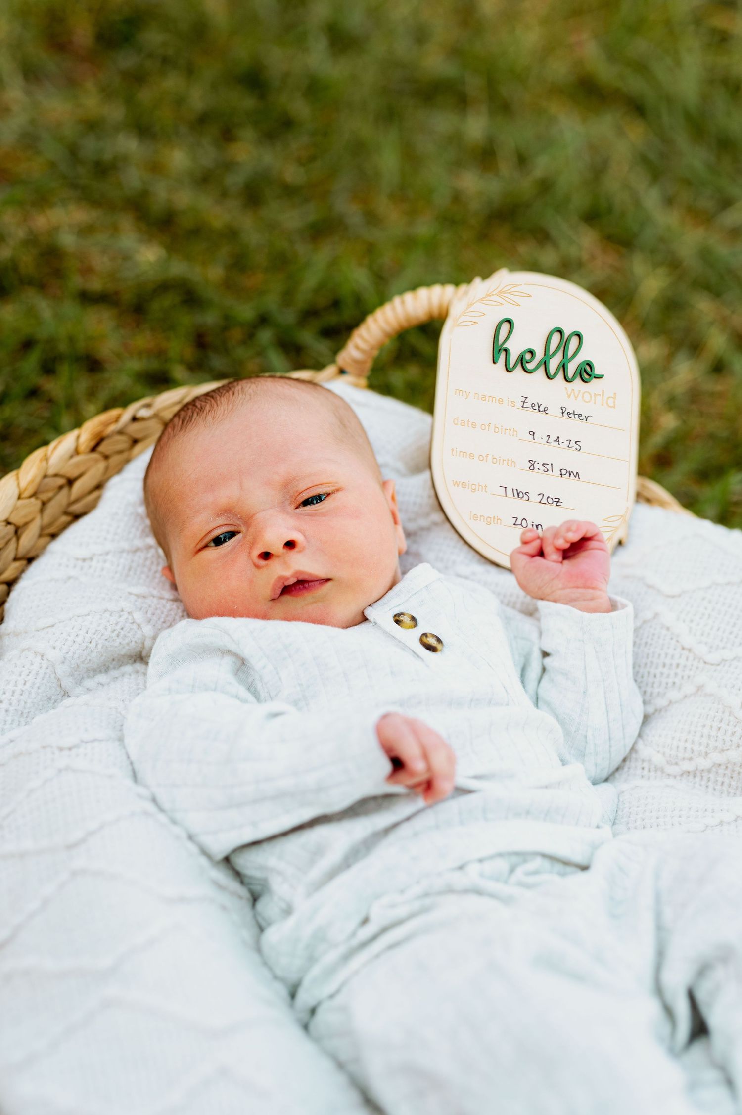 Baby in white outfit lying on blanket outdoors with milestone marker showing name and date.