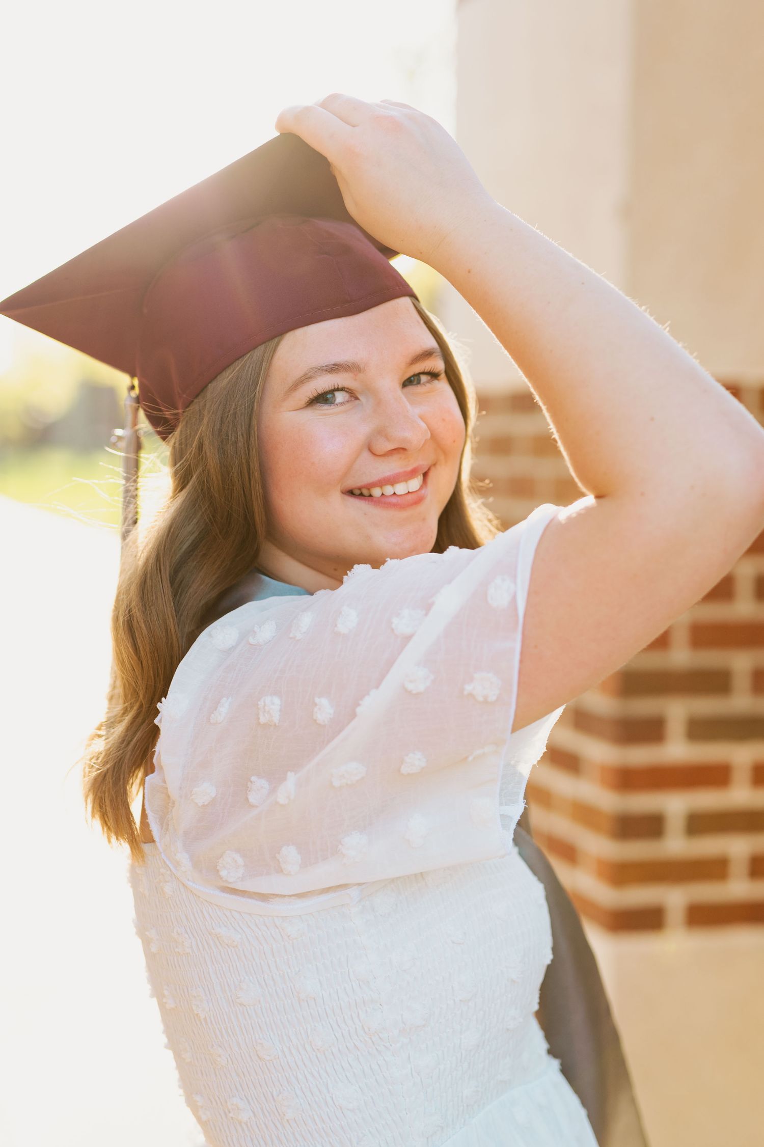 A graduate adjusts cap while posing in white dress during sunset photoshoot.