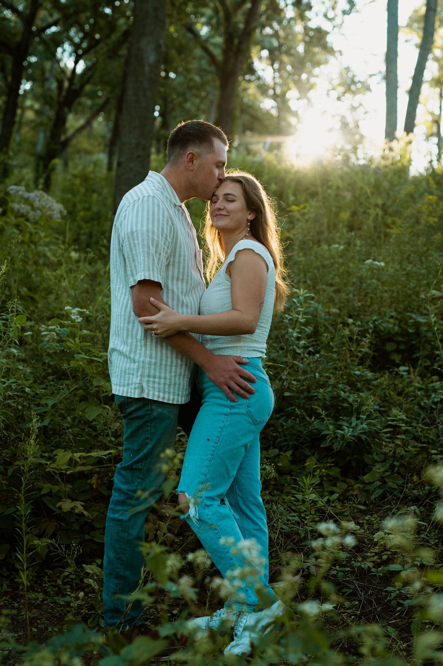 Couple in casual attire sharing tender moment in sunlit wooded area.