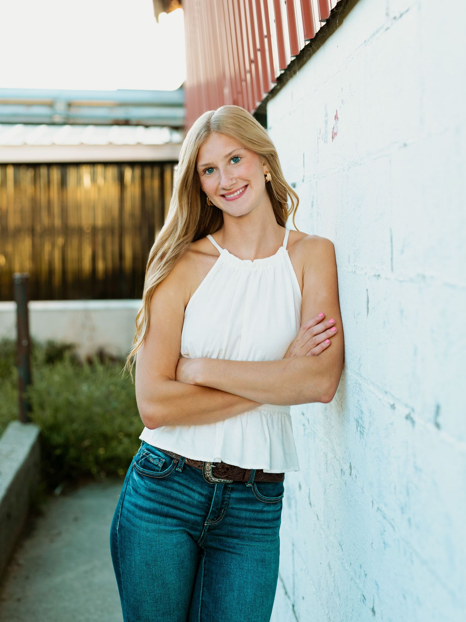 A young woman in blue jeans and white top poses against a white wall in a casual outdoor photoshoot.