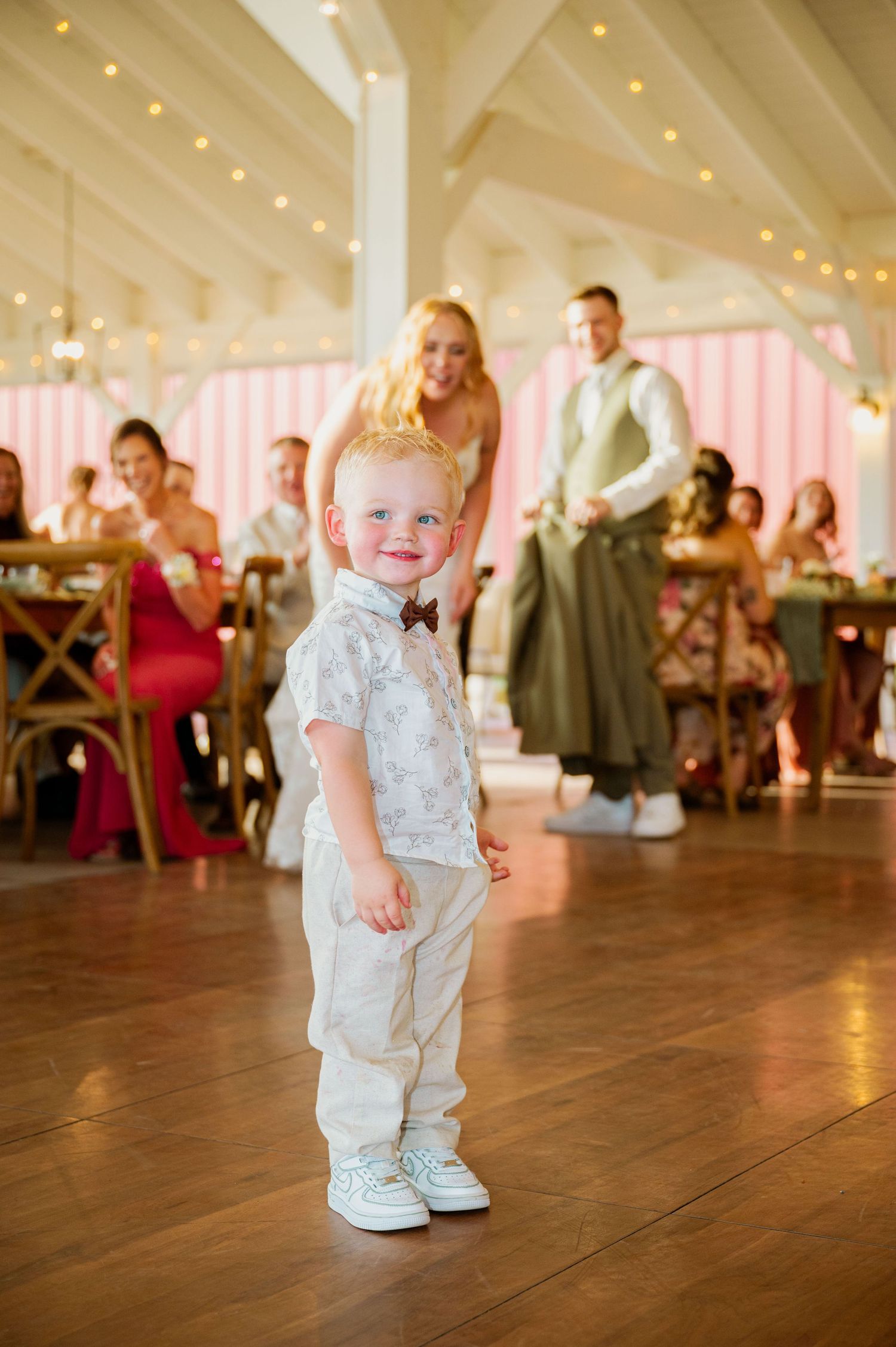 Adorable toddler in formal attire dances on wooden floor under twinkling lights at wedding reception.