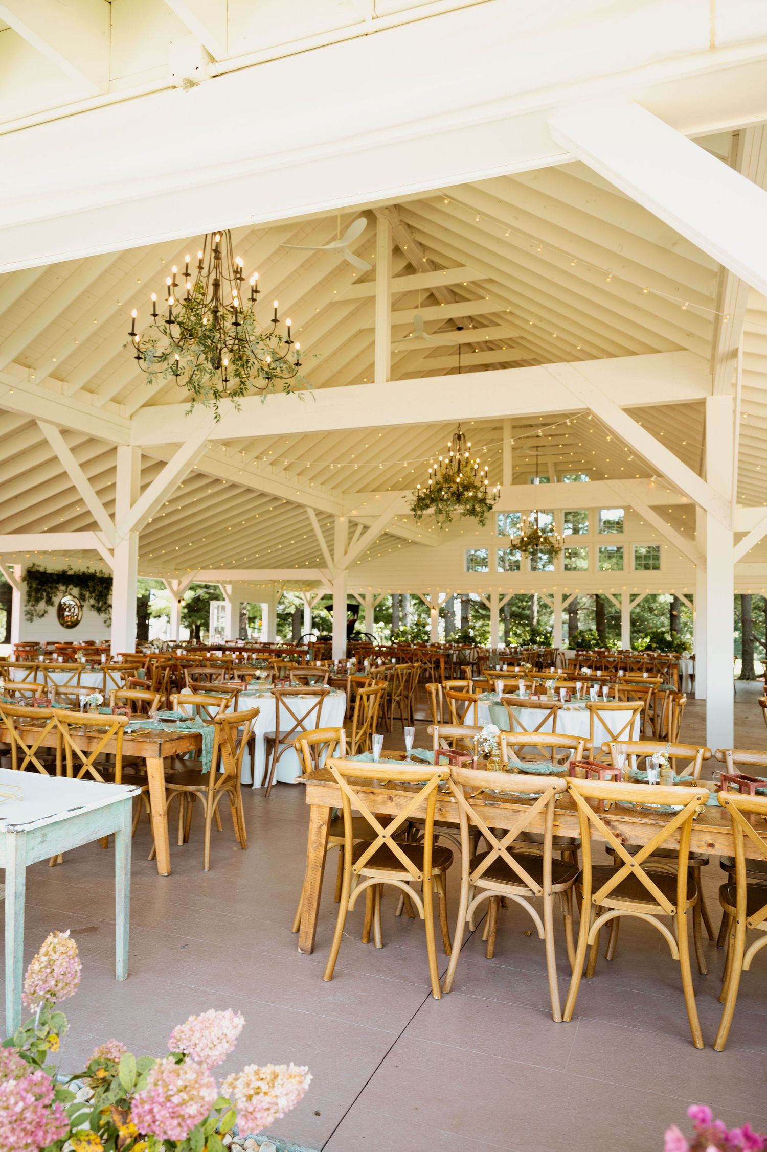 Elegant event space with cross-back chairs, chandeliers, and pink floral accents under a white pavilion roof.