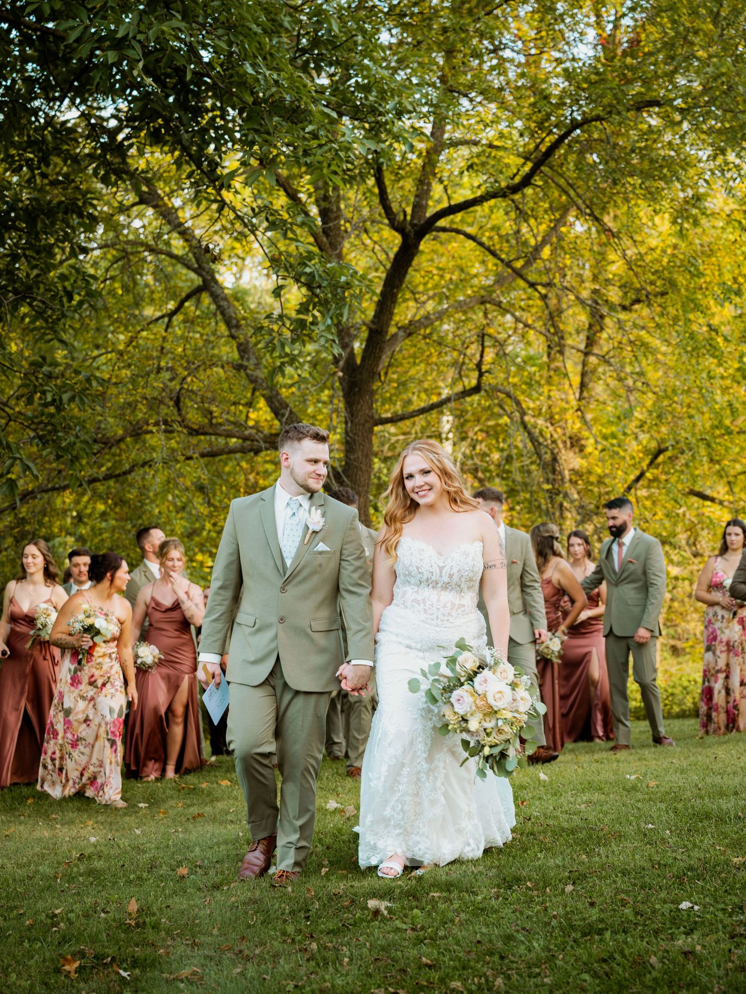Wedding party in sage green and dusty rose attire poses outdoors under golden trees.