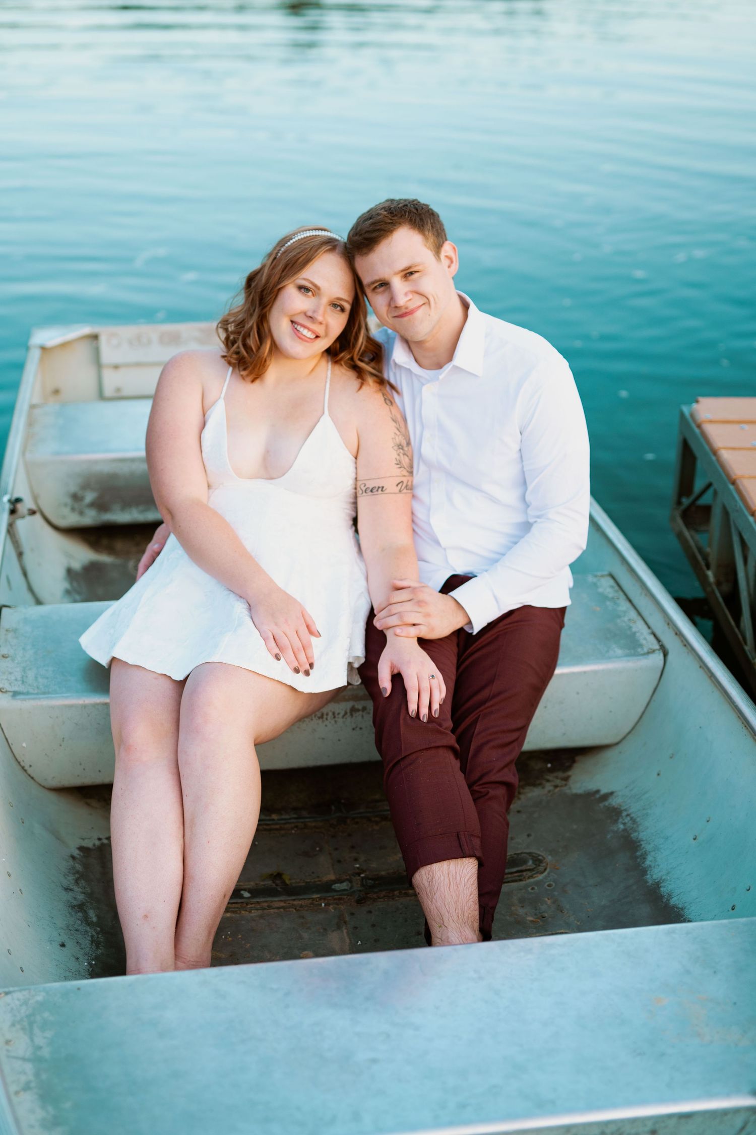 A romantic couple sits together in a rowboat on tranquil blue water during a photography session.