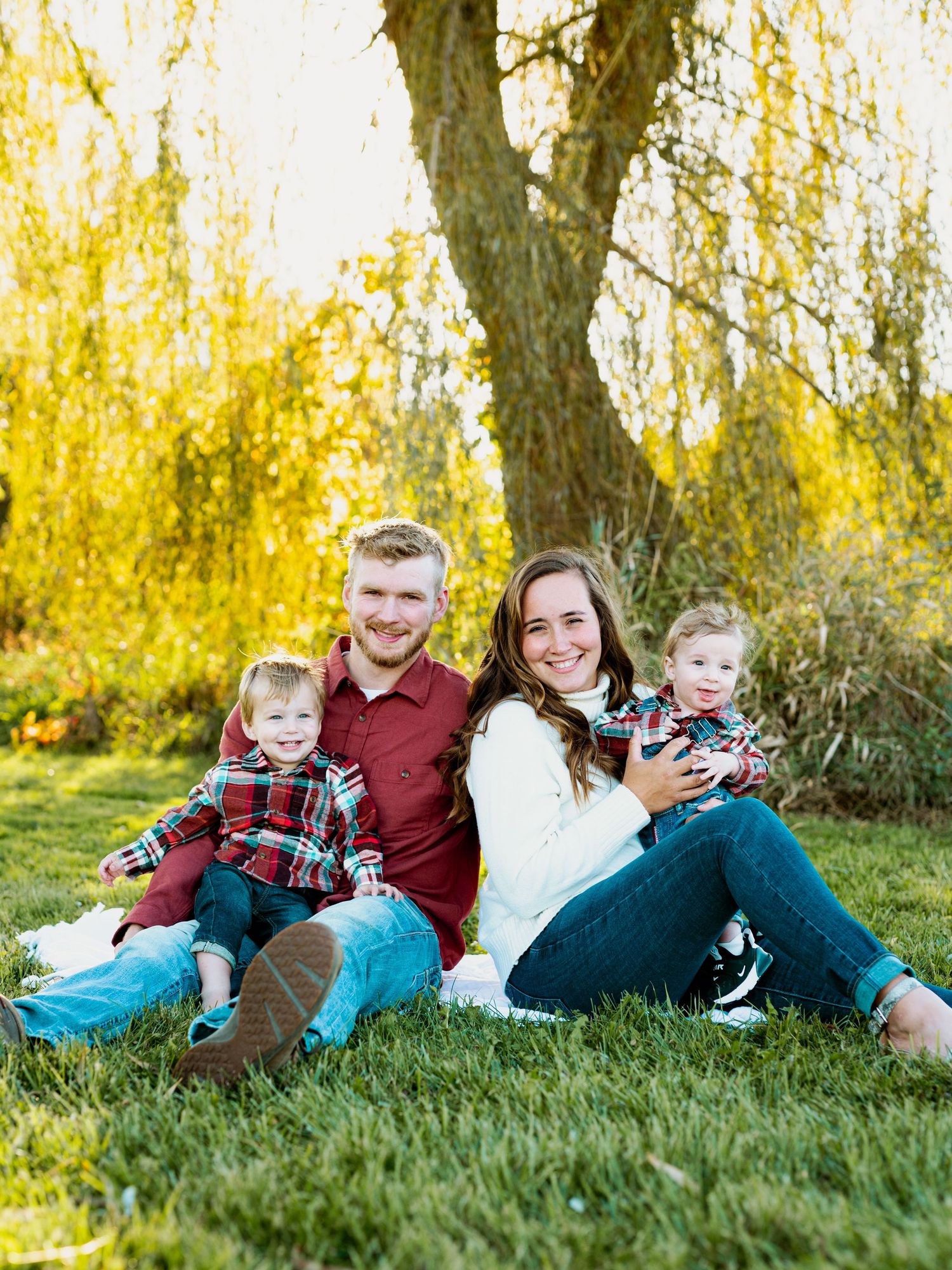 Family sits together on grass under a willow tree with golden autumn foliage in the background.
