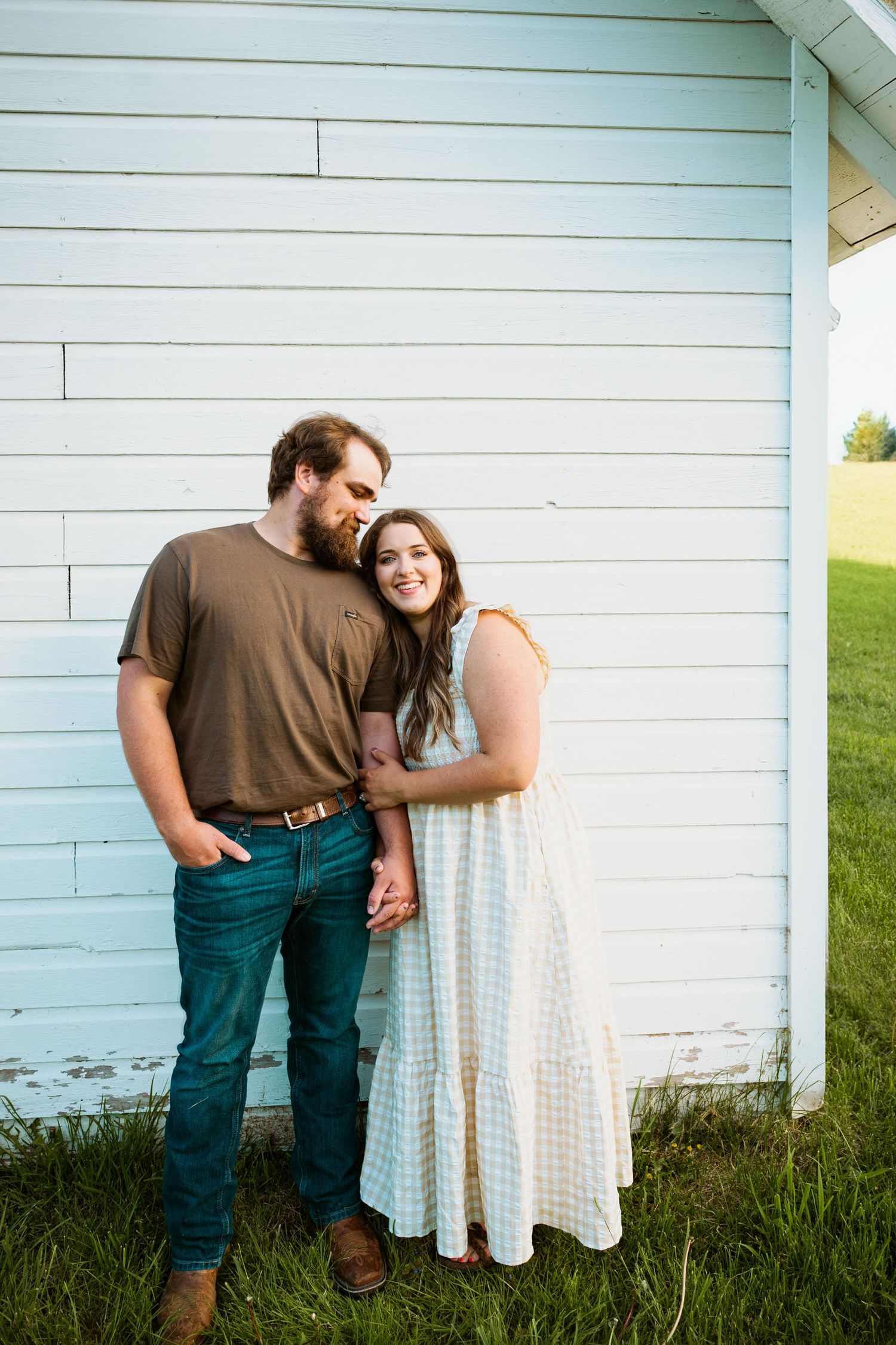 A couple shares a tender moment against a white wooden wall, with her in a flowing dress and him in brown and teal.