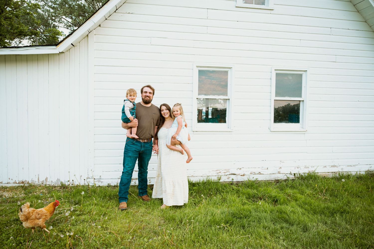 A white farmhouse serves as backdrop for a family photoshoot on spacious green lawn.
