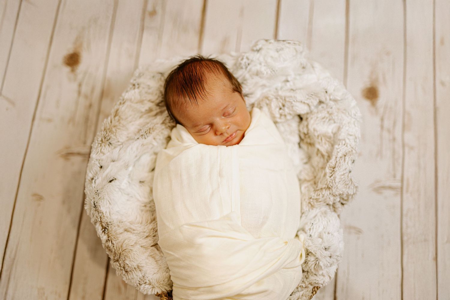 Newborn baby swaddled in white fabric resting on plush white material against wooden floor.