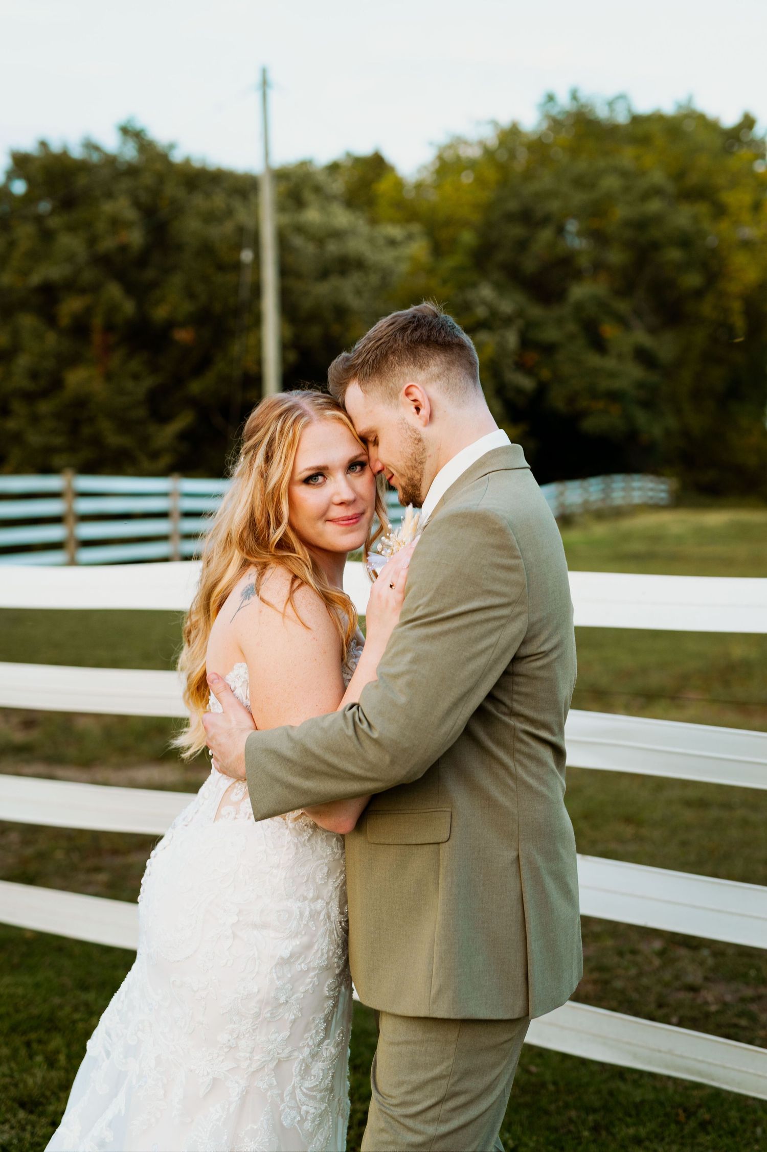 Romantic couple portrait against white fence during sunset at rustic farm venue.