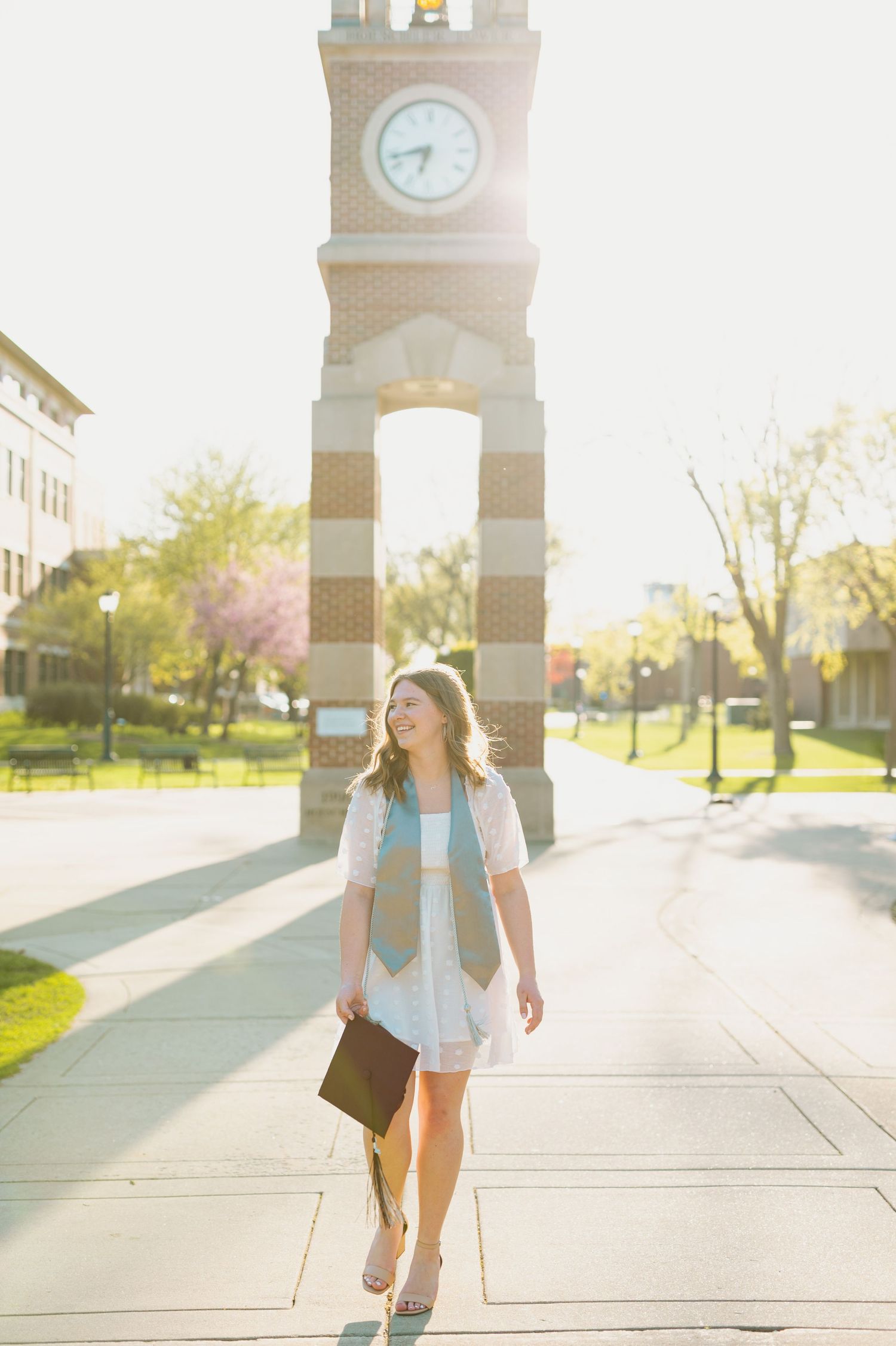 A graduate in white dress walks past a university clock tower in golden sunlight.