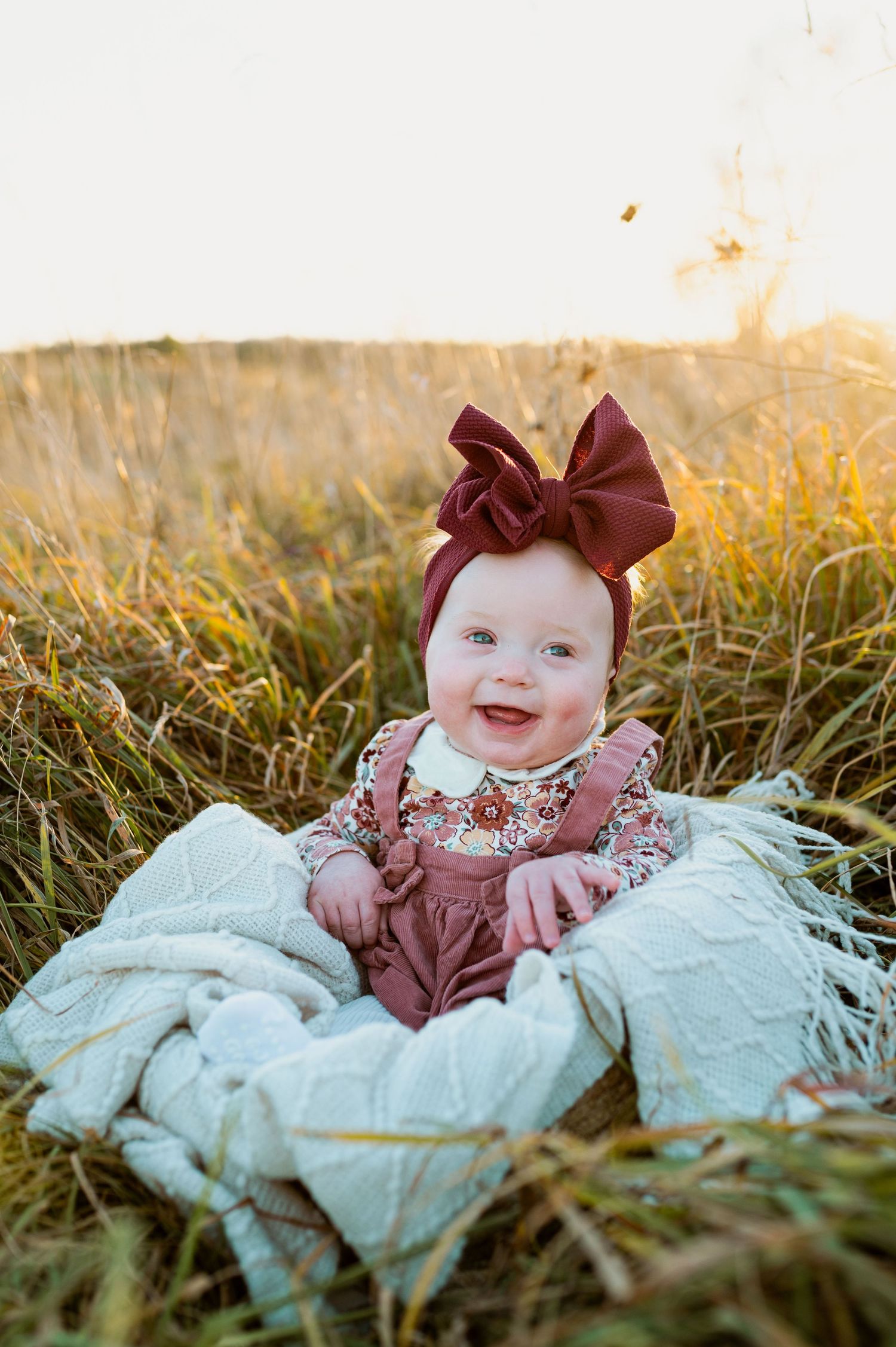 A small baby wearing a burgundy bow and outfit lies on white fabric in autumn grass at sunset.