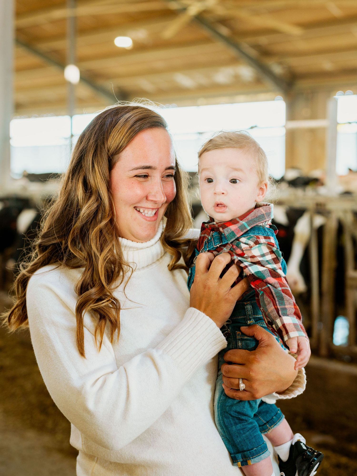 A woman in a white turtleneck sweater shares tender moments with a baby in denim overalls in a rustic barn setting.