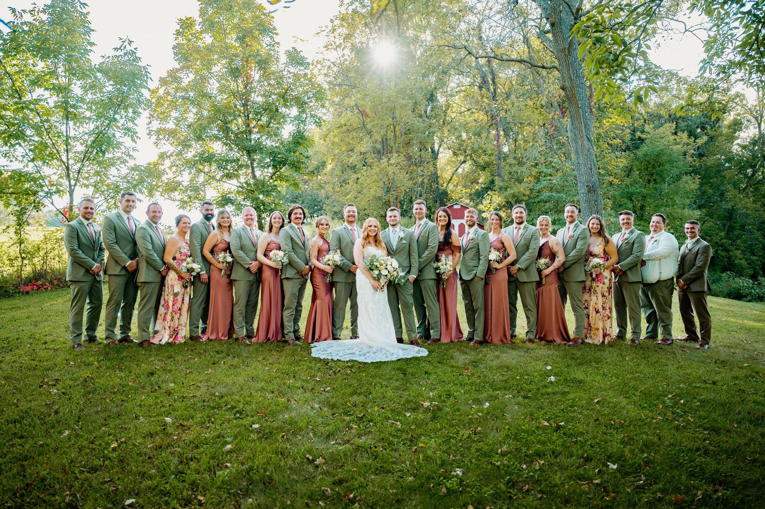 Wedding party in burgundy and tan attire poses together on a grassy hillside at sunset.