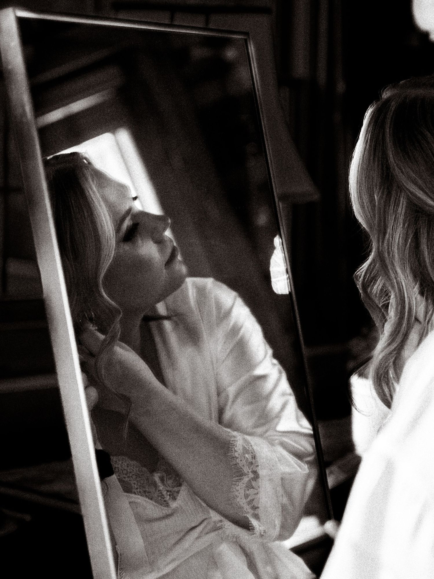 Bride in white dress looking through window blinds creating dramatic lighting and shadows.
